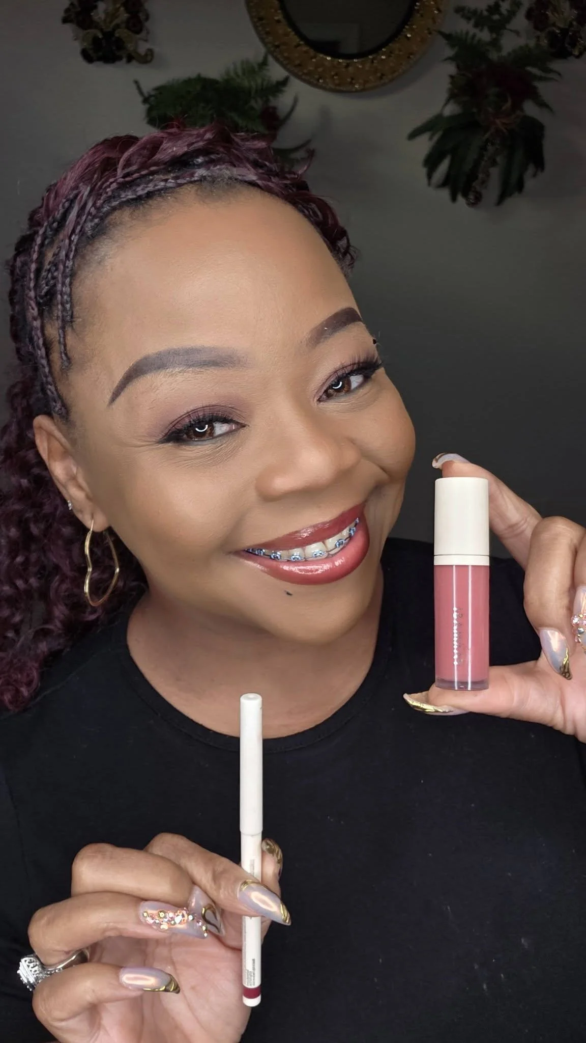 A woman smiling and holding a lip gloss and a lip liner, showing her makeup and braces.