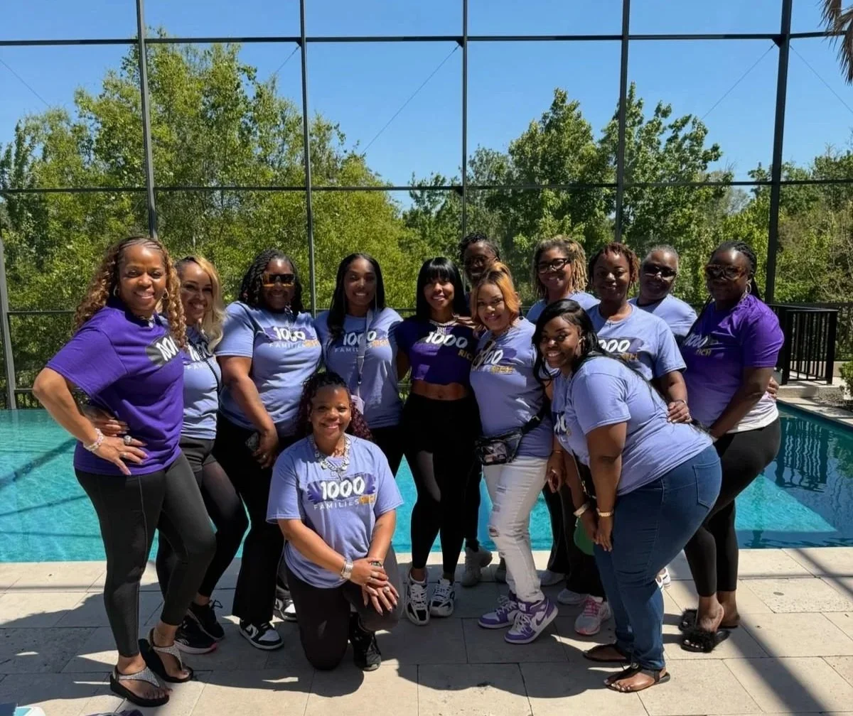 Group of women standing together near a swimming pool, wearing matching purple and blue T-shirts with '1000 Families' written on them, outdoors on a sunny day with trees and a blue sky in the background.