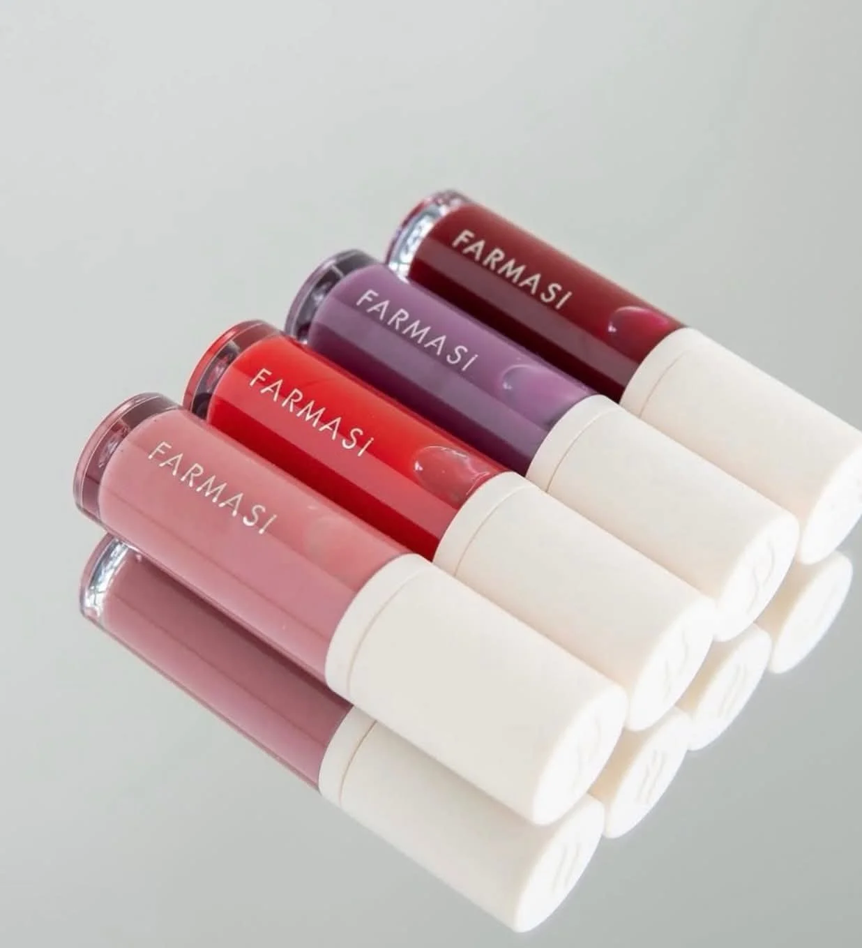 Five tubes of lip gloss labeled Farmasi in various shades ranging from pink to purple and red, laid on a reflective surface.