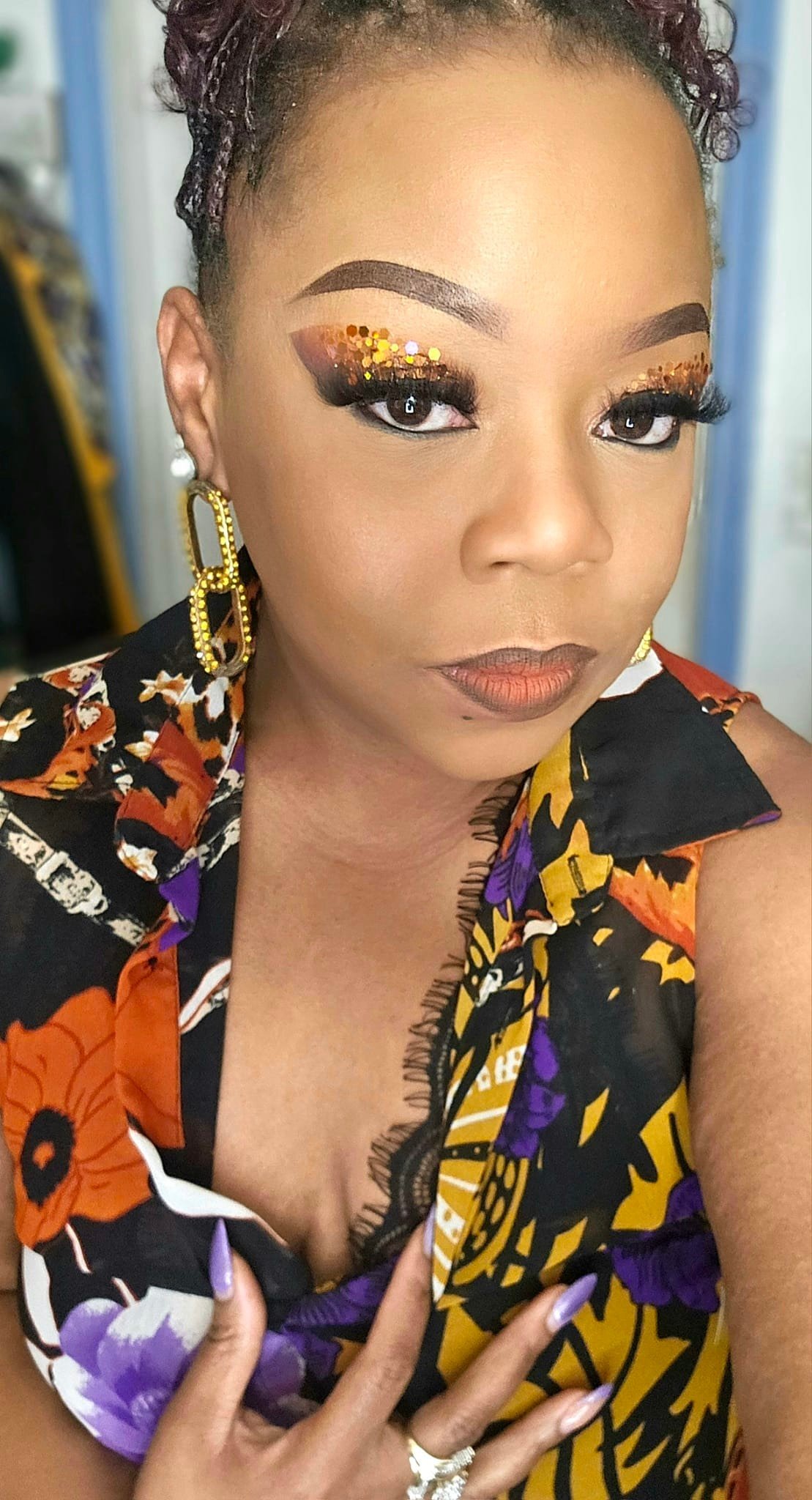 A woman with curly purple hair styled in an updo, wearing bold makeup with glitter eyeshadow, false eyelashes, and brown lipstick. She is dressed in a colorful, floral-patterned black, purple, orange, and yellow sleeveless top with lace details, large yellow earrings, and is showcasing a purple manicure and a ring.