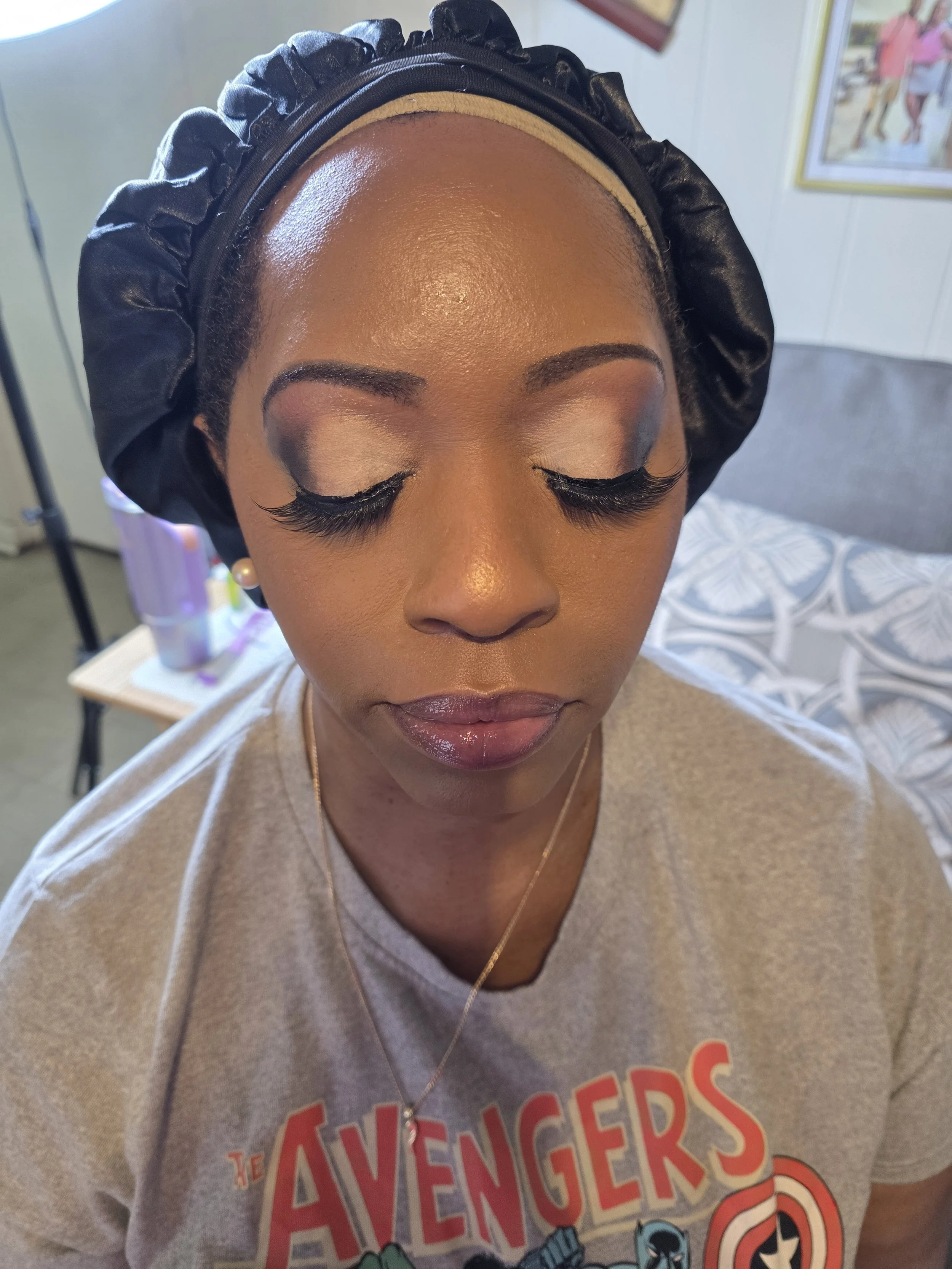 Close-up of a woman with her eyes closed, wearing makeup, a black satin bonnet, pearl earrings, and an Avengers T-shirt.