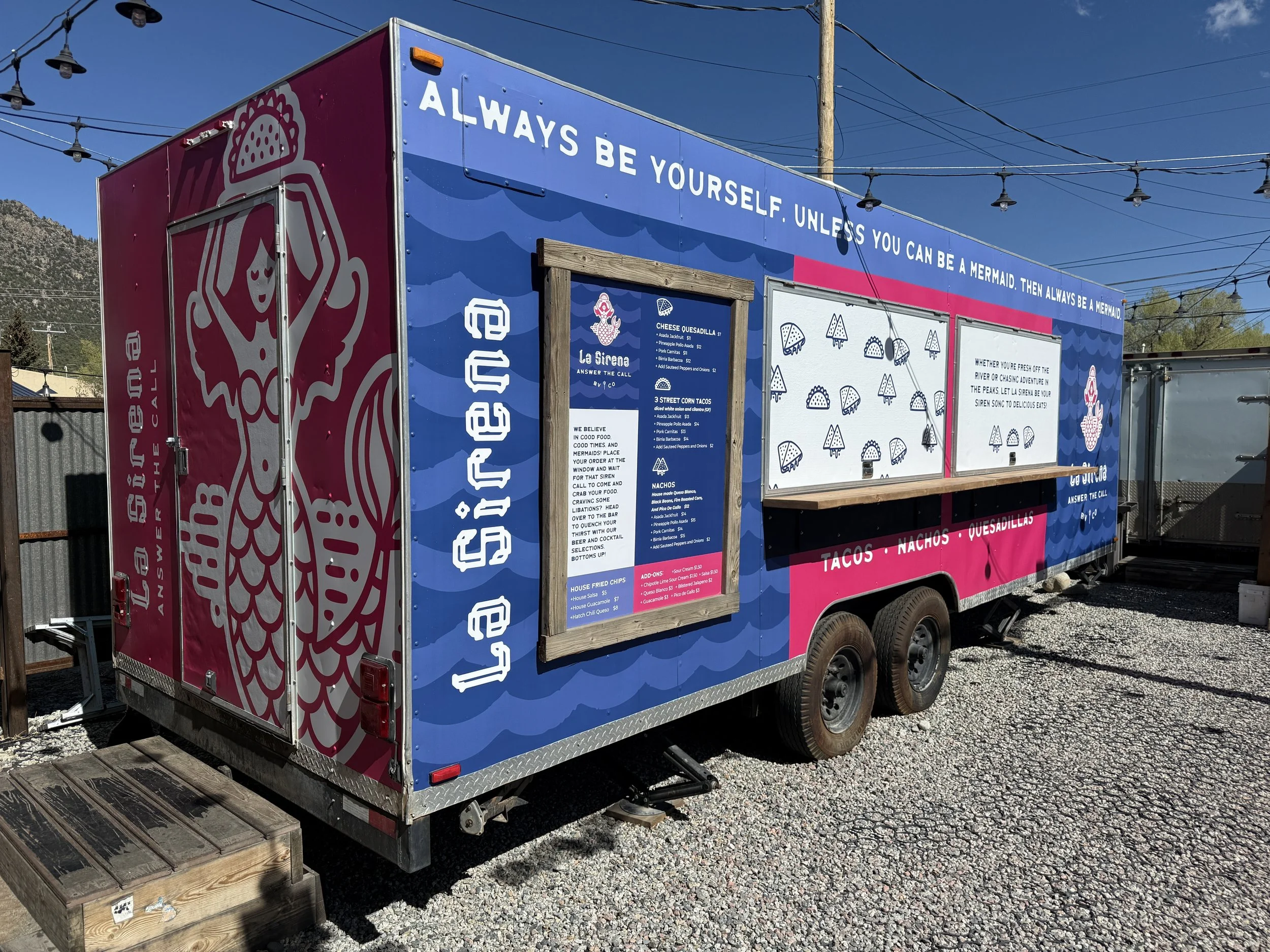 Colorful food truck with a blue and pink design and a mermaid theme. The truck has menu boards on the side and quote at the top saying, "Always be yourself. Unless you can be a mermaid. Then always be a mermaid." The food truck serves tacos, nachos, 