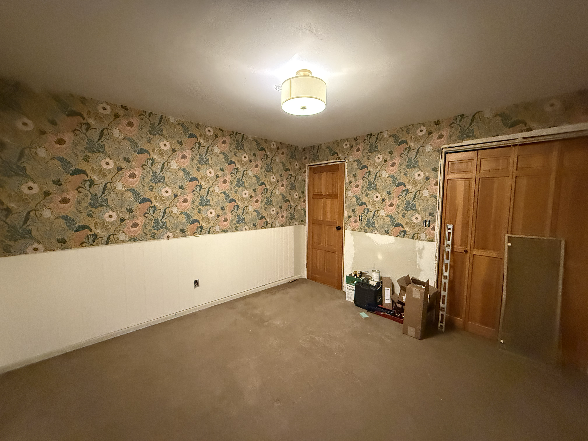 Empty room with floral wallpaper on the upper walls, white lower wall paneling, a wooden door, built-in wooden closet, and various boxes and tools near the closet.