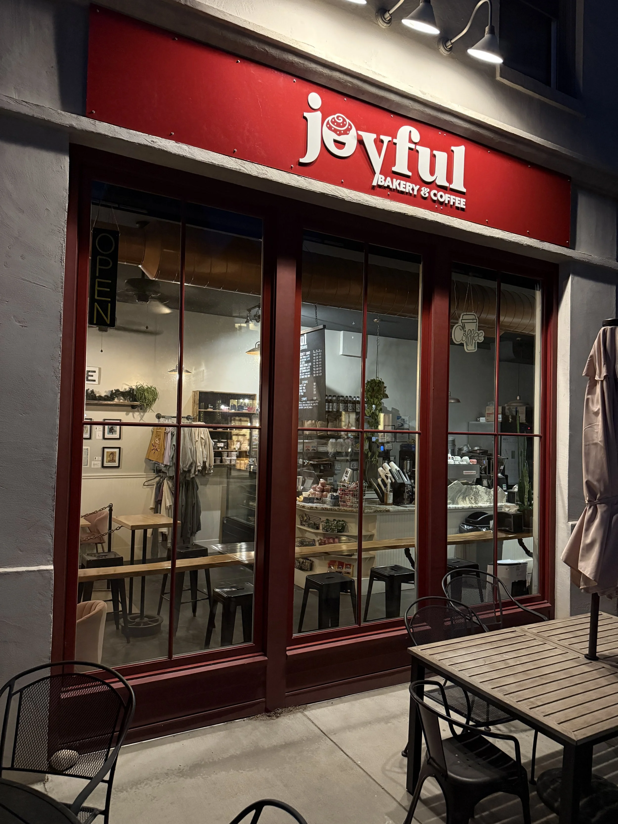 The exterior of a bakery and coffee shop named Joyful with a red sign and large windows showing the interior, which has a counter, seating, and decorations.