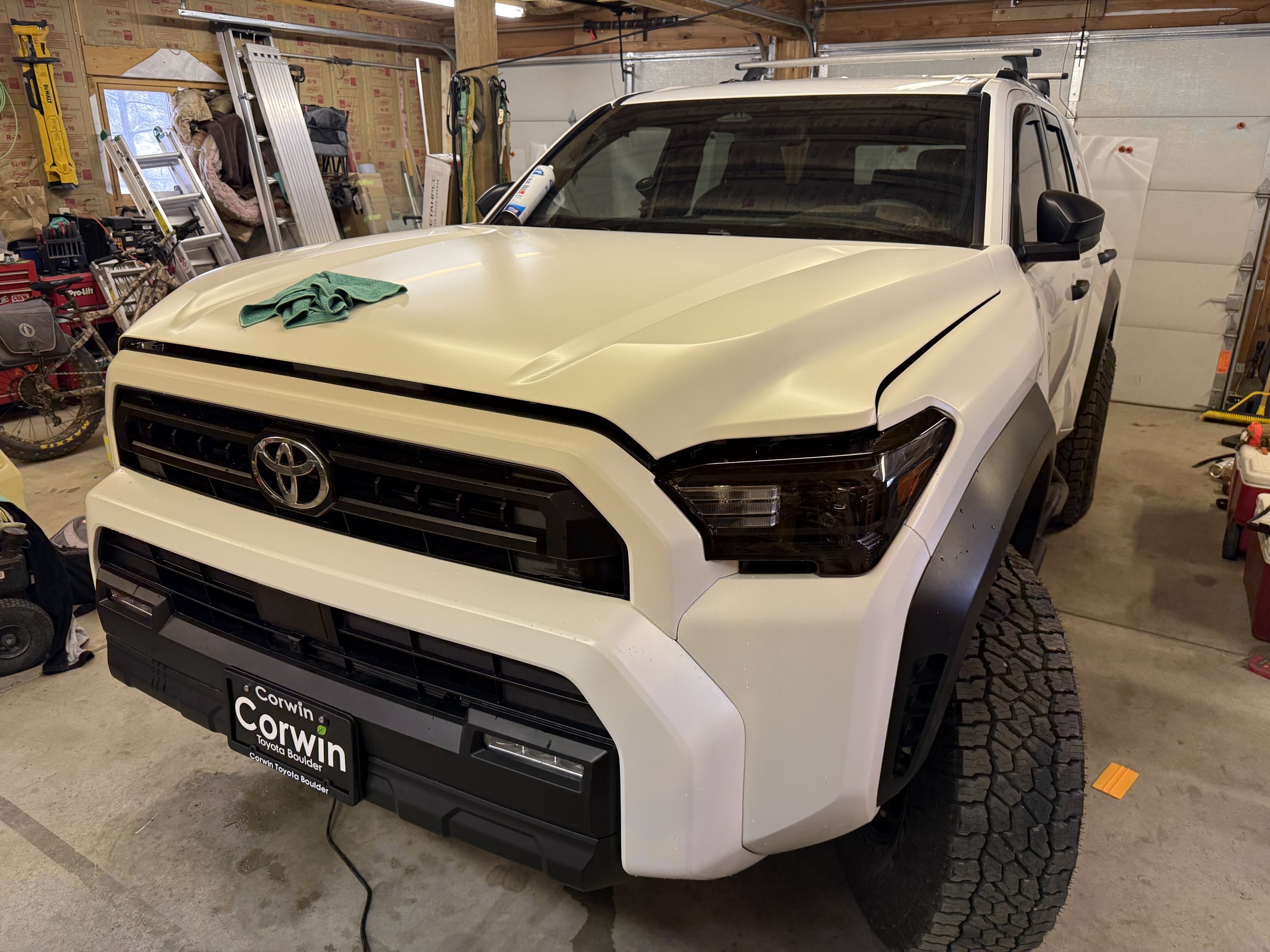 A white Toyota truck with black accents parked inside a garage or workshop. The front hood has a green cloth on it, and the vehicle has large, off-road tires. The background shows tools, ladders, and other equipment.