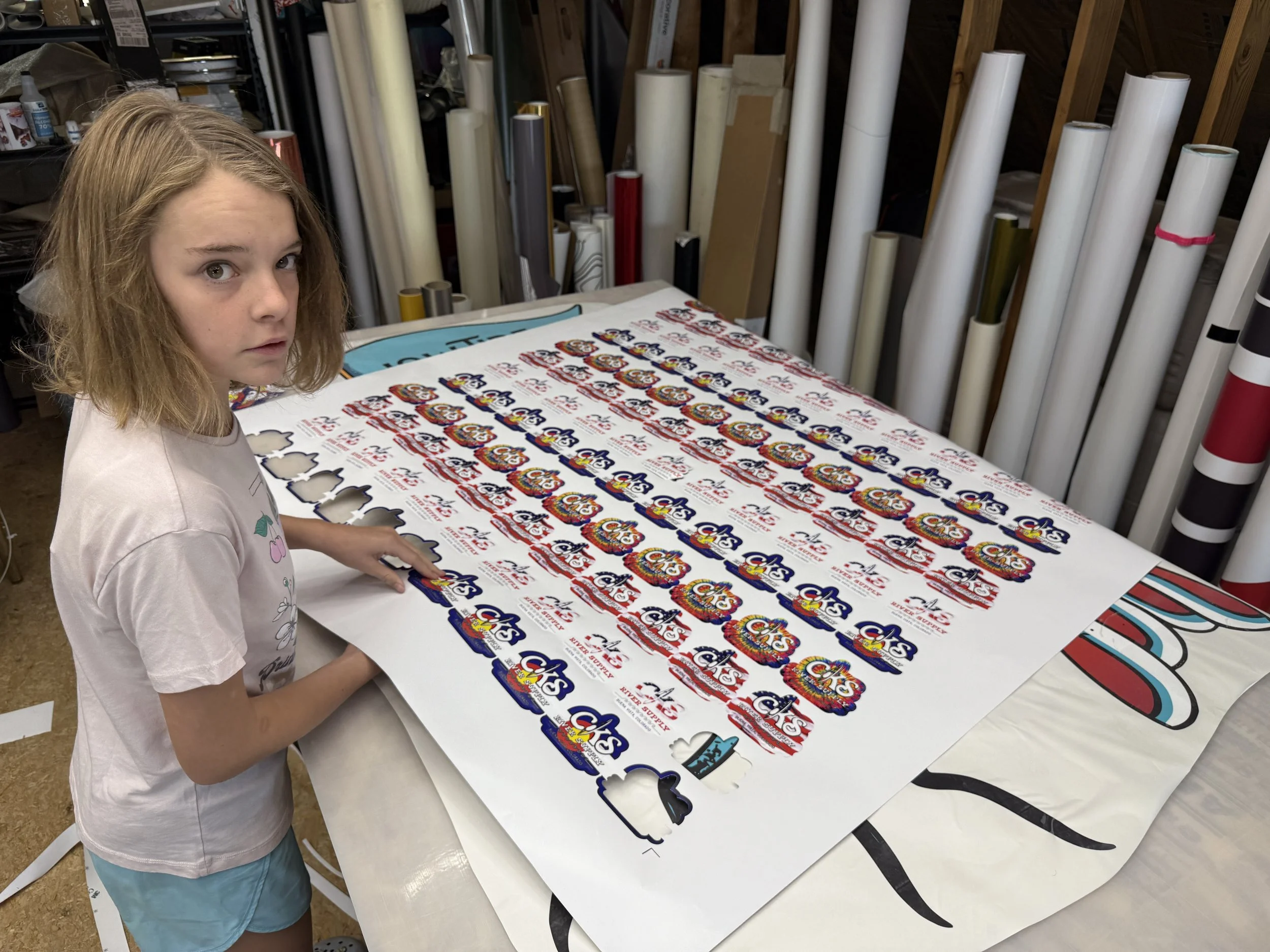 A girl with shoulder-length light brown hair looks at the camera while working on a large decal sheet with colorful logos and text, surrounded by rolls of vinyl and other materials in a workshop.