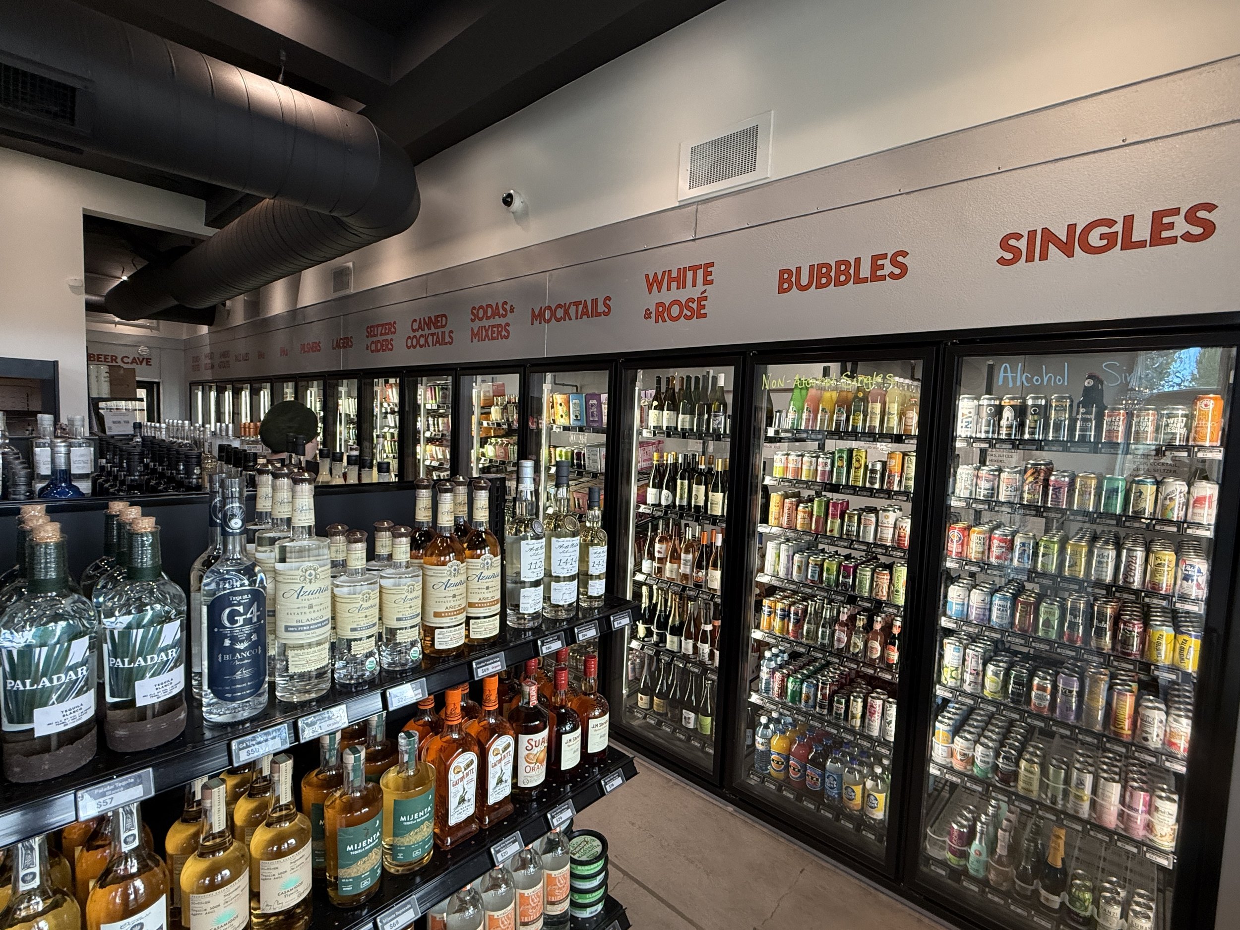 View of a liquor store aisle with refrigerated beer and alcohol, shelves with various bottles of whiskey and spirits, and a overhead sign listing product categories such as beers, ciders, mixers, and singles.
