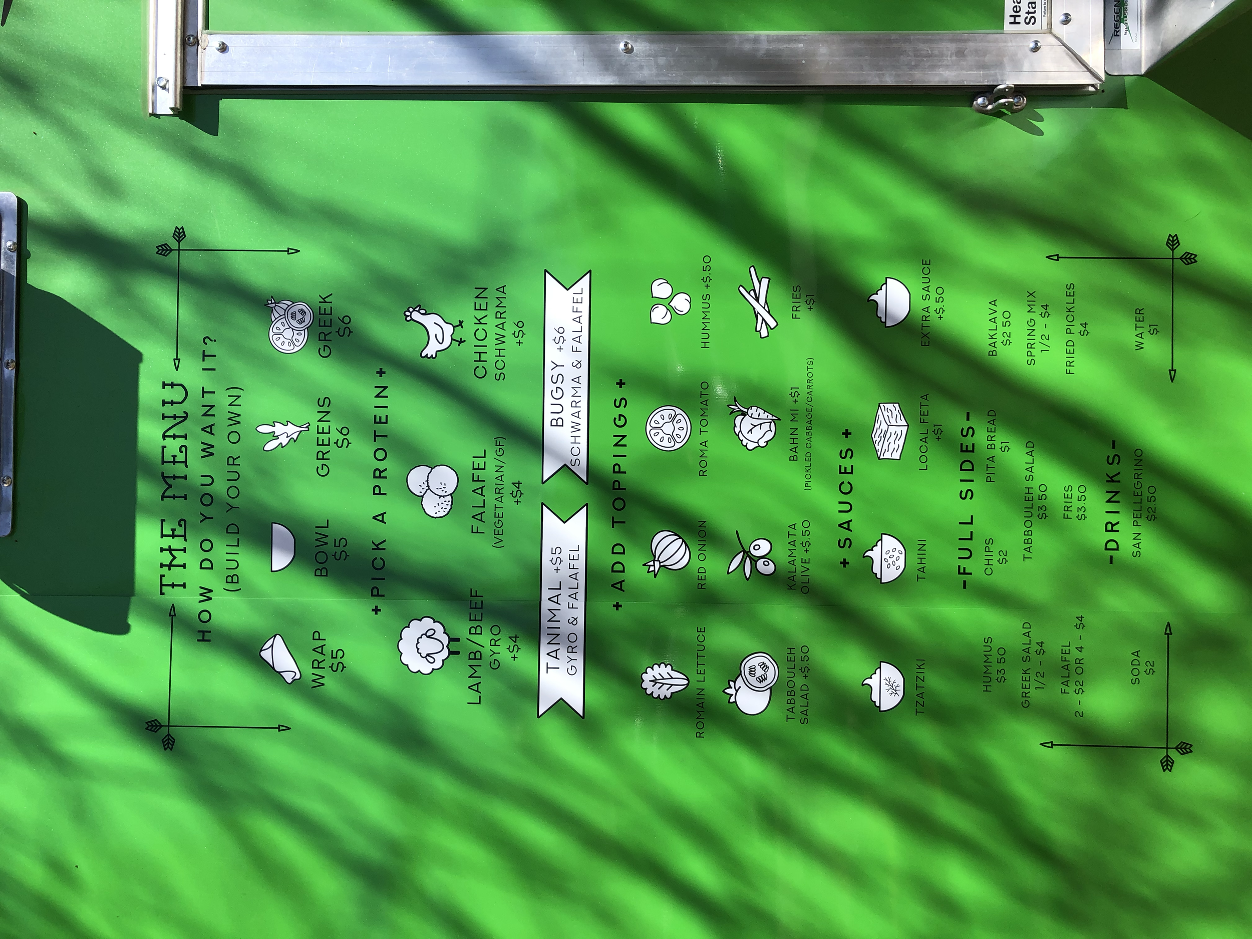 Green menu board with food options written in black and white, including salads, wraps, falafel, gyros, noodle bowls, and drinks.