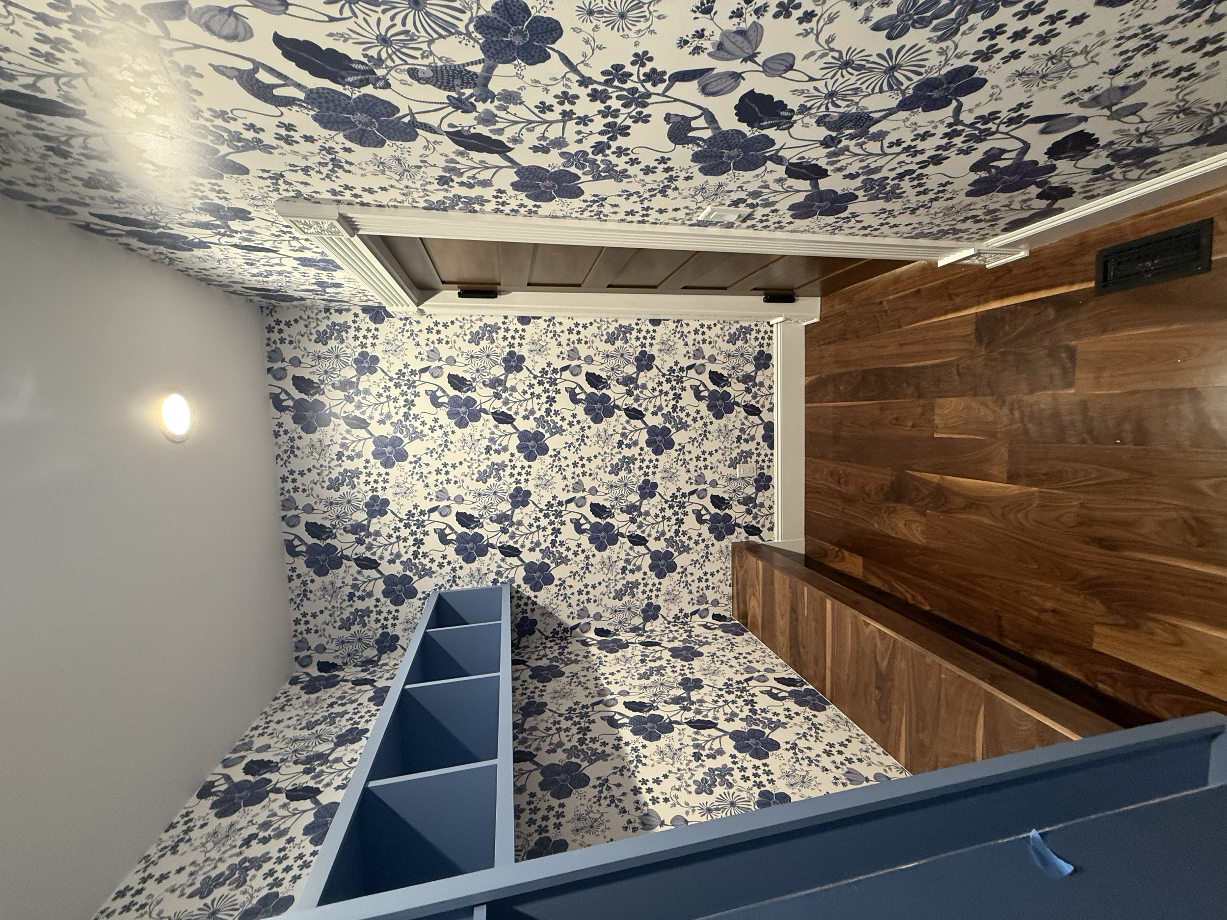 A small room with floral blue and white wallpaper, a wooden door, and a wooden bench. The ceiling has a recessed light fixture, and there is a built-in shelving unit painted blue.