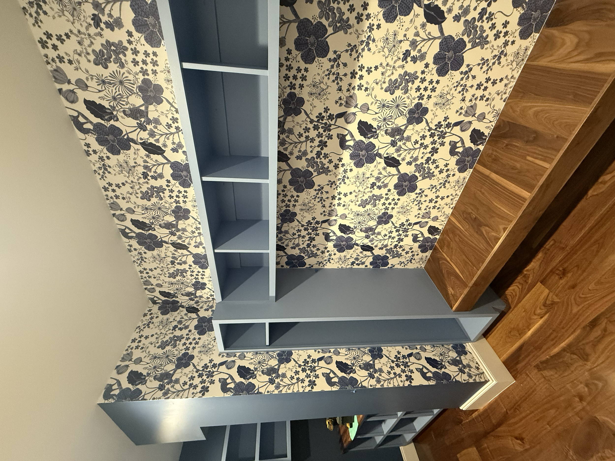 Room with floral-patterned wallpaper, blue bookshelves, and wood flooring.