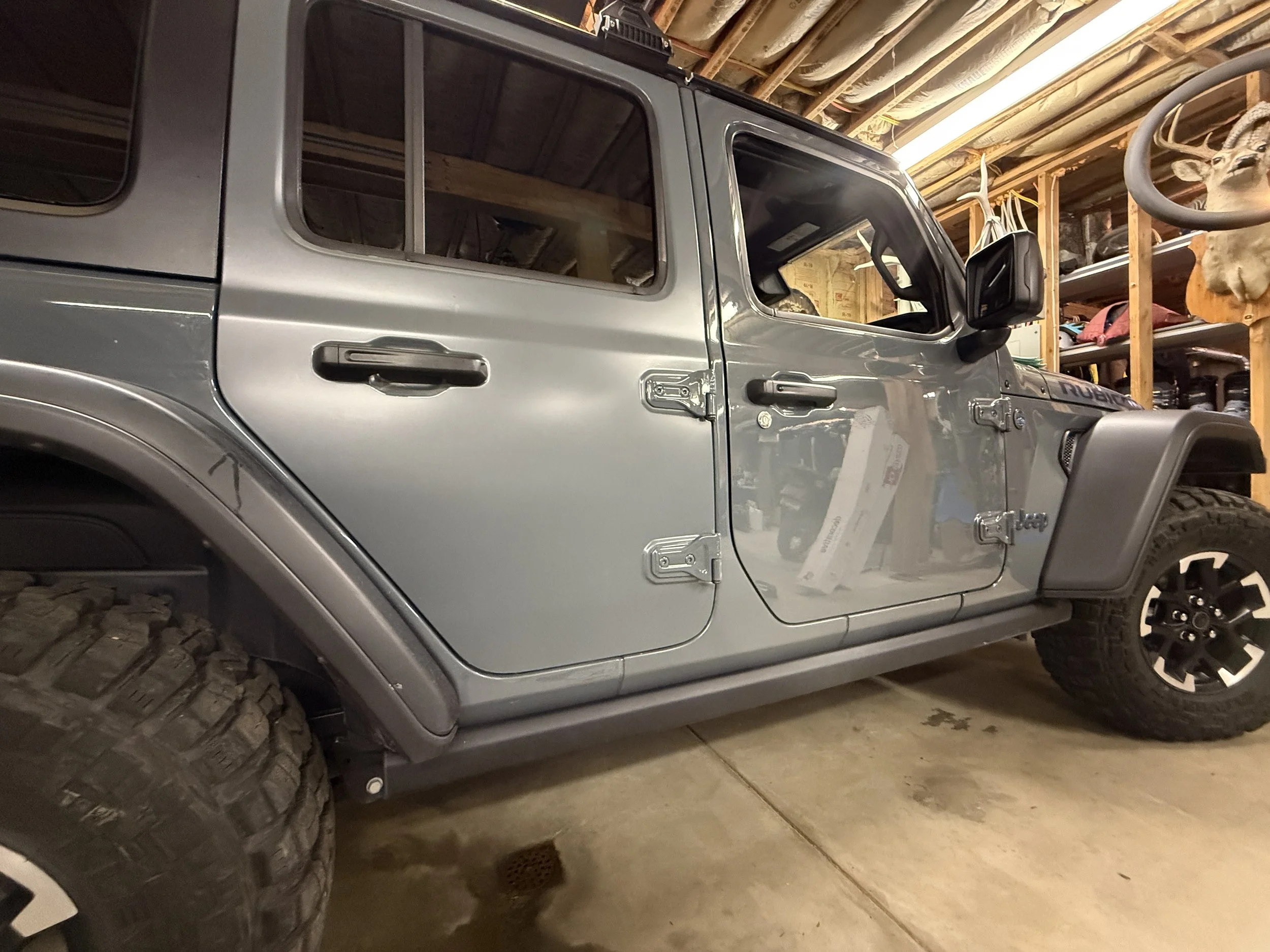 Gray Jeep Rubicon with four doors parked in a garage, partially assembled, with parts and shelves on the wall.