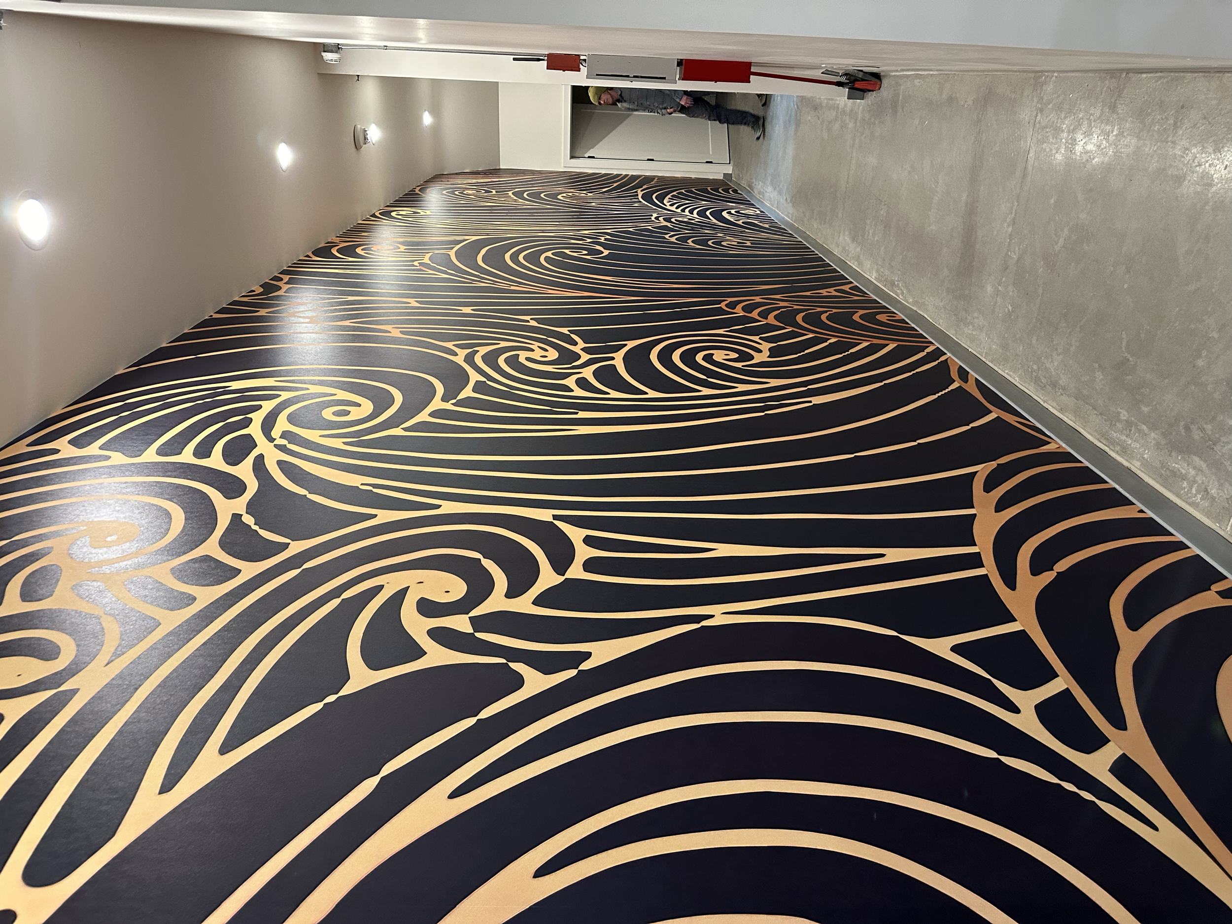 A hallway with a black and orange swirling patterned carpet, plain gray wall on the right, and a white ceiling with recessed lighting.