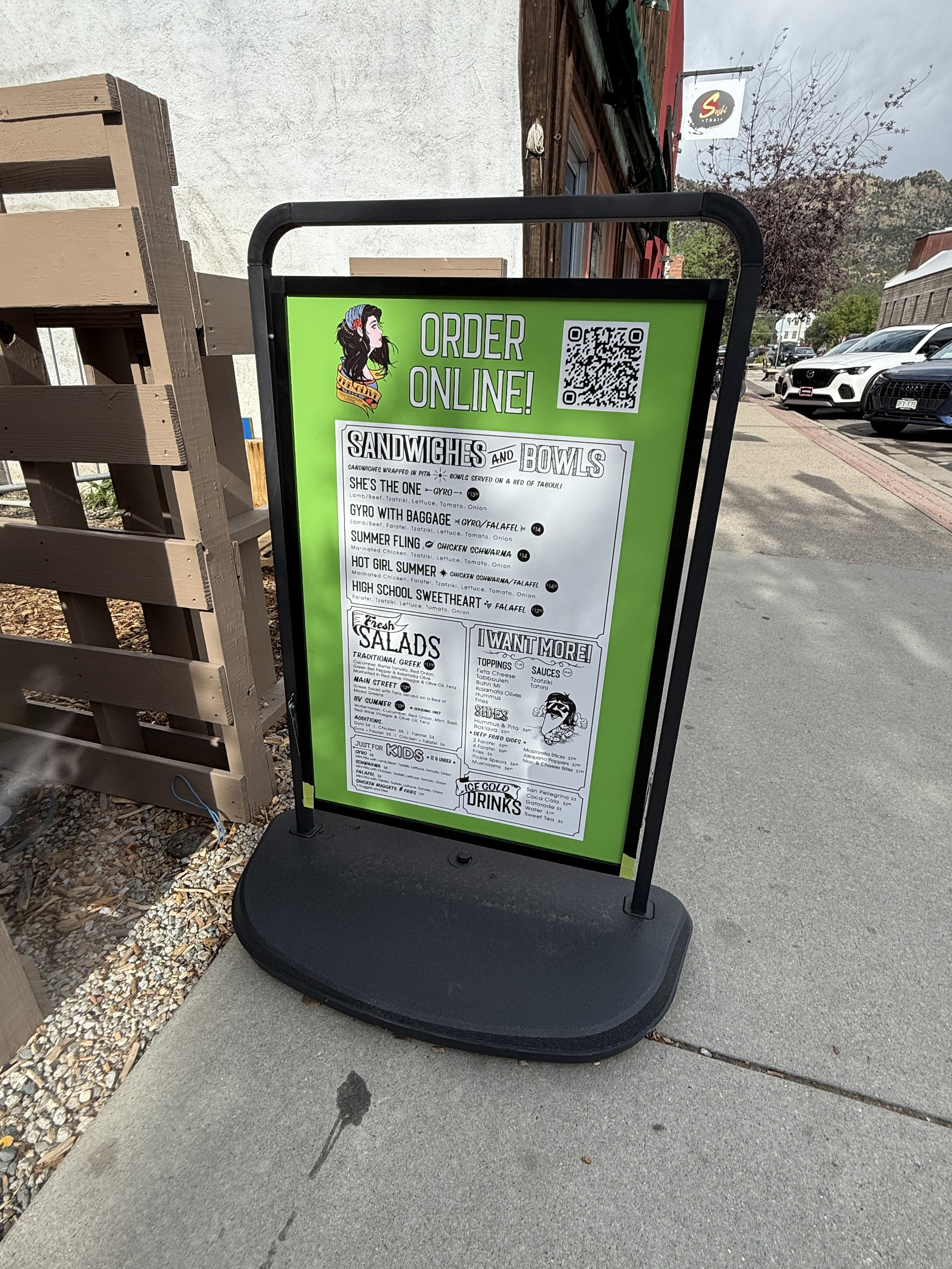 A green and white outdoor restaurant menu sign promoting online ordering with a QR code. The menu lists sandwiches, bowls, salads, and drinks, with some illustrations and headings.