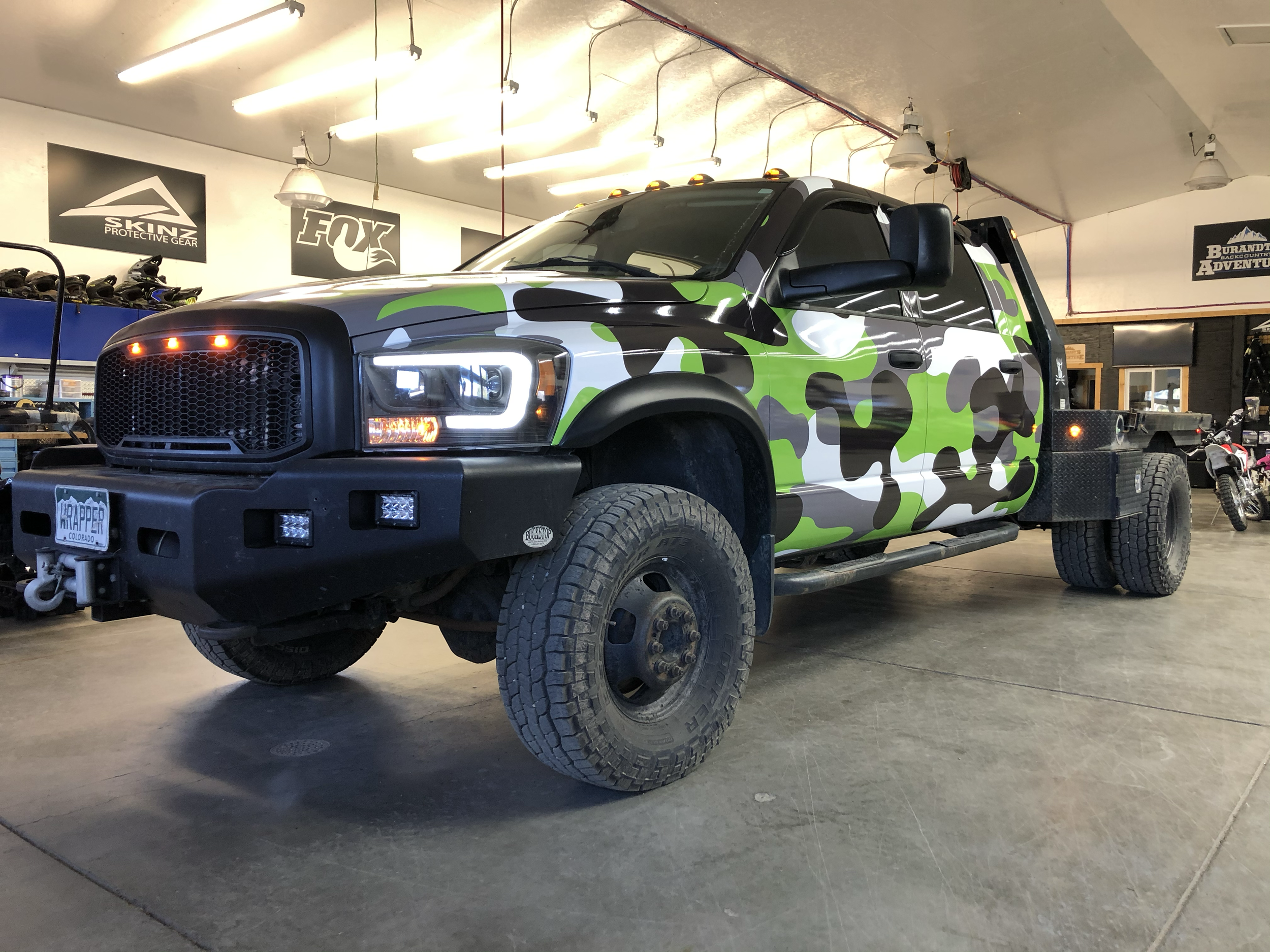 A lifted, heavy-duty truck with a green, black, white, and gray camouflage paint job inside a showroom or garage, with equipment and banners in the background.