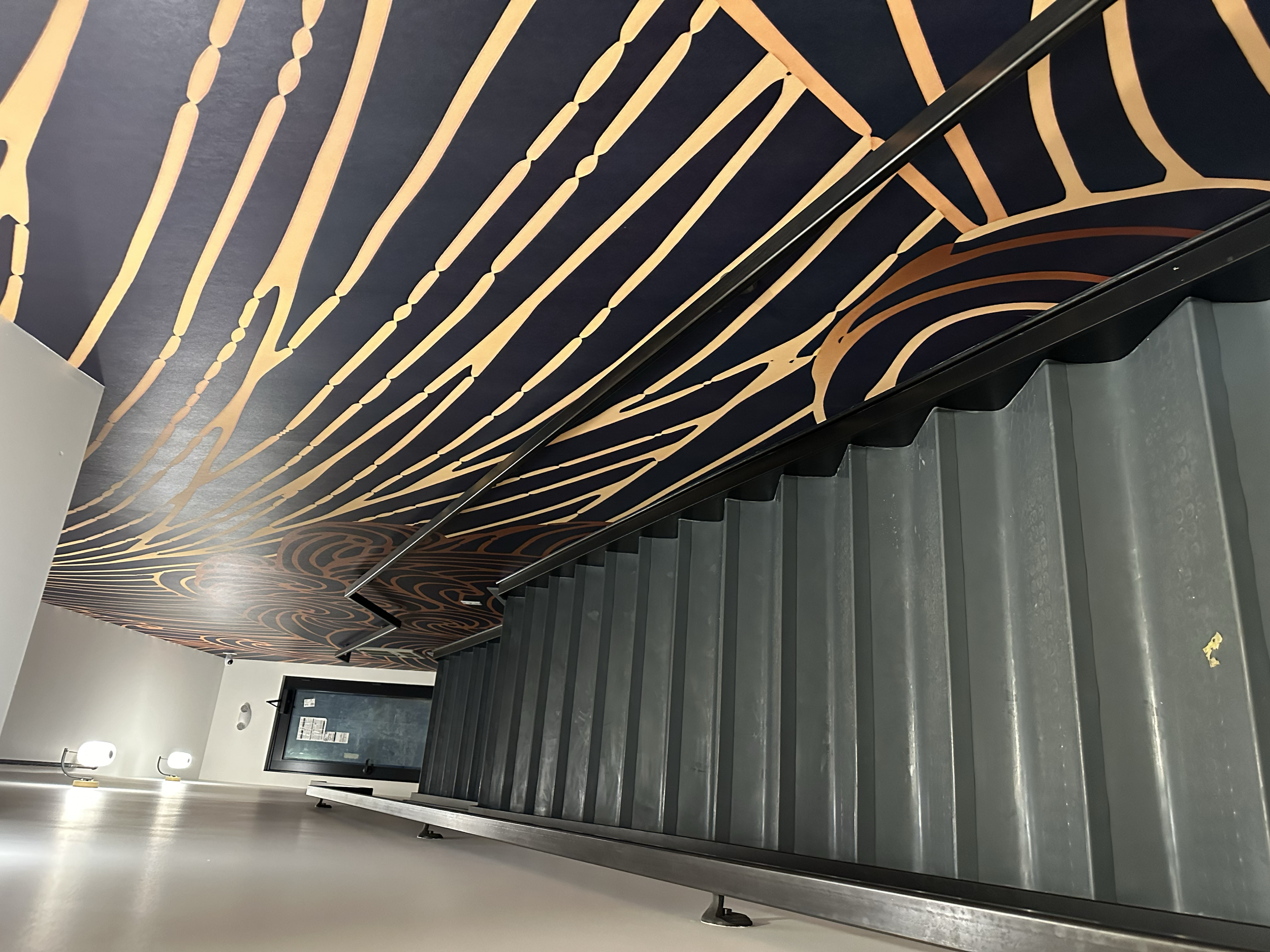 Interior view of a stairwell with black and gold patterned wallpaper on the wall and stair steps leading downward.