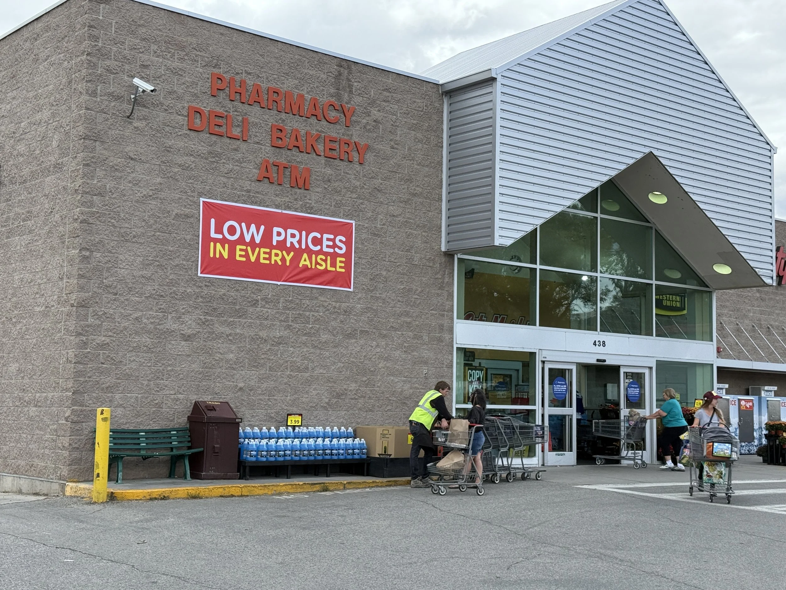People entering a supermarket with shopping carts outside the entrance. A sign on the wall advertises low prices in every aisle. The store has a brick wall and a glass front entrance.