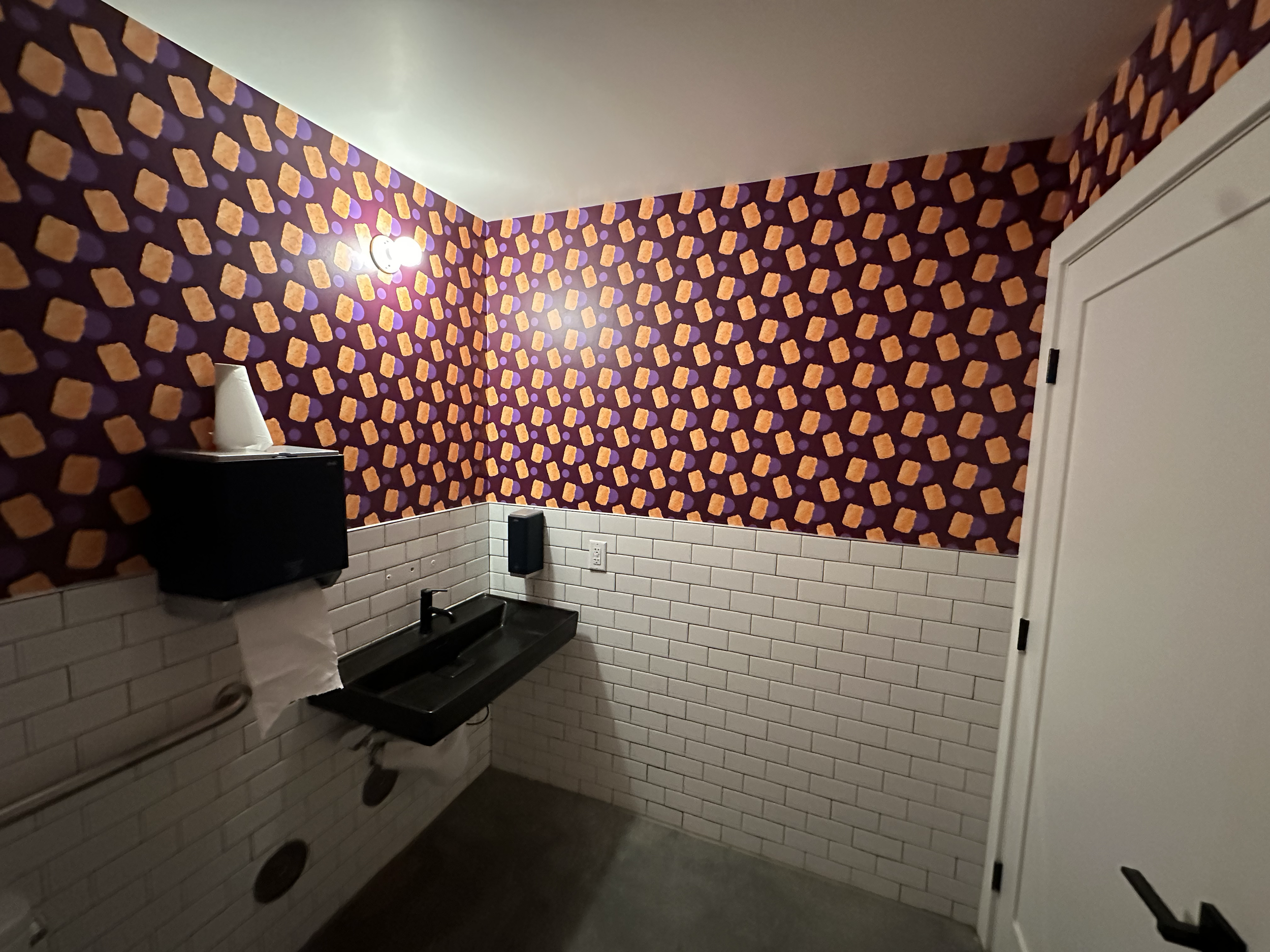 A bathroom with purple and orange patterned wallpaper, white subway tile halfway up the wall, a black sink, a paper towel dispenser, and a closed white door.