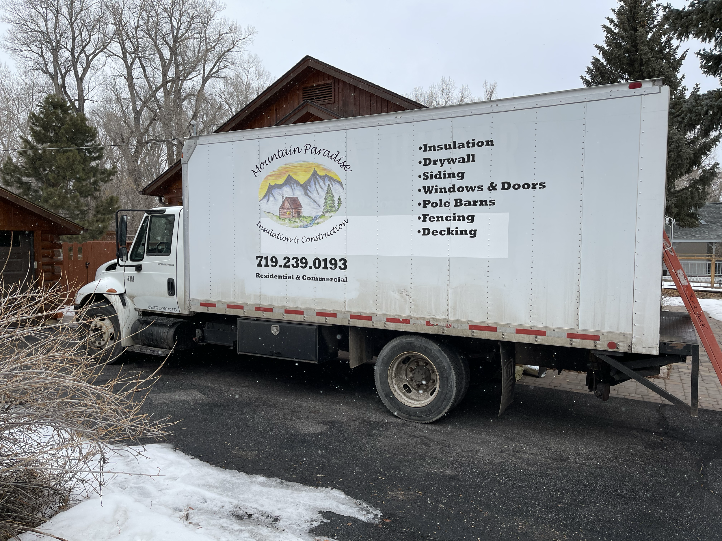 A white truck parked on a driveway with a business advertisement on its side for Mountain Paradise Insulation & Construction, including a mountain and cabin logo, a phone number, and a list of services such as insulation, drywall, siding, windows and