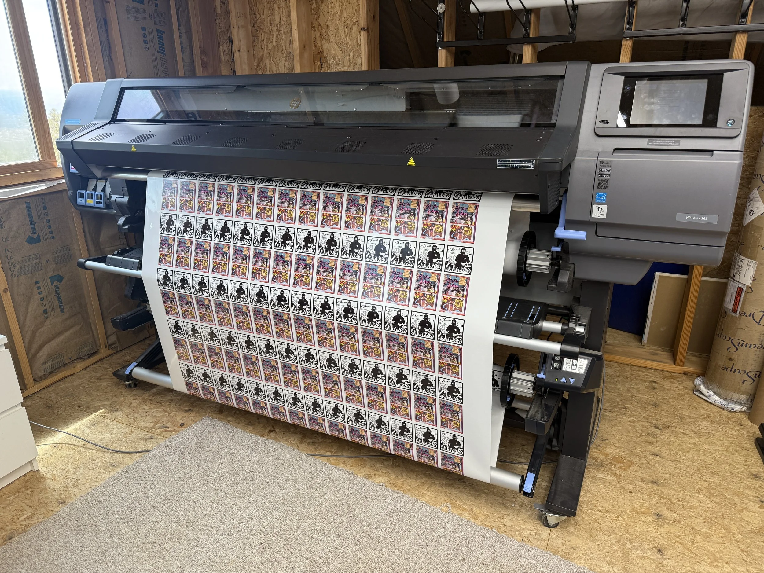 Large printer producing multiple sheets of colorful stickers with black silhouettes and vibrant graphic backgrounds in a wooden-themed room.