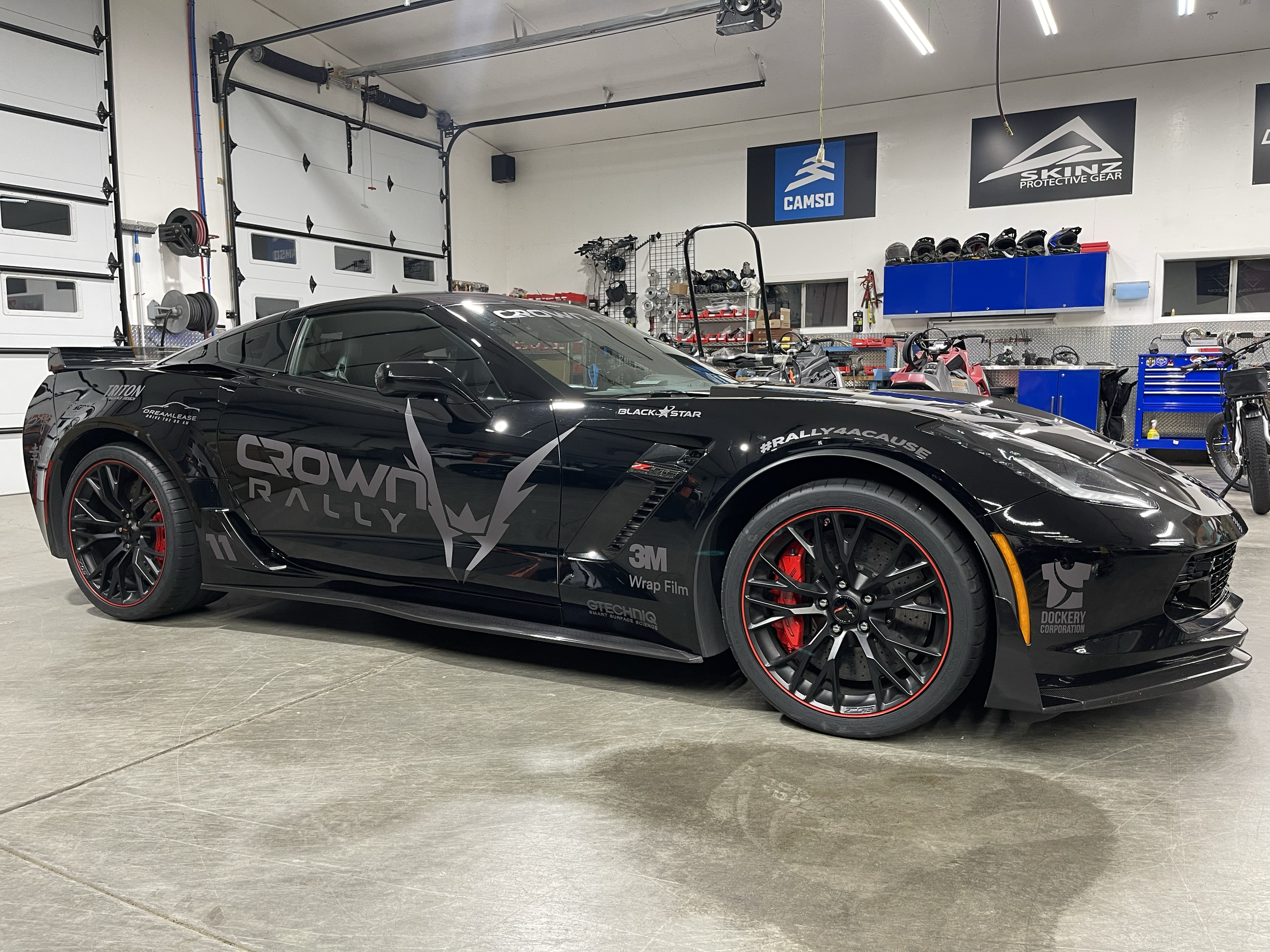 A black Chevrolet Corvette with Crown Rally branding and red brake calipers parked inside a garage with tools, helmets, and banners on the walls.