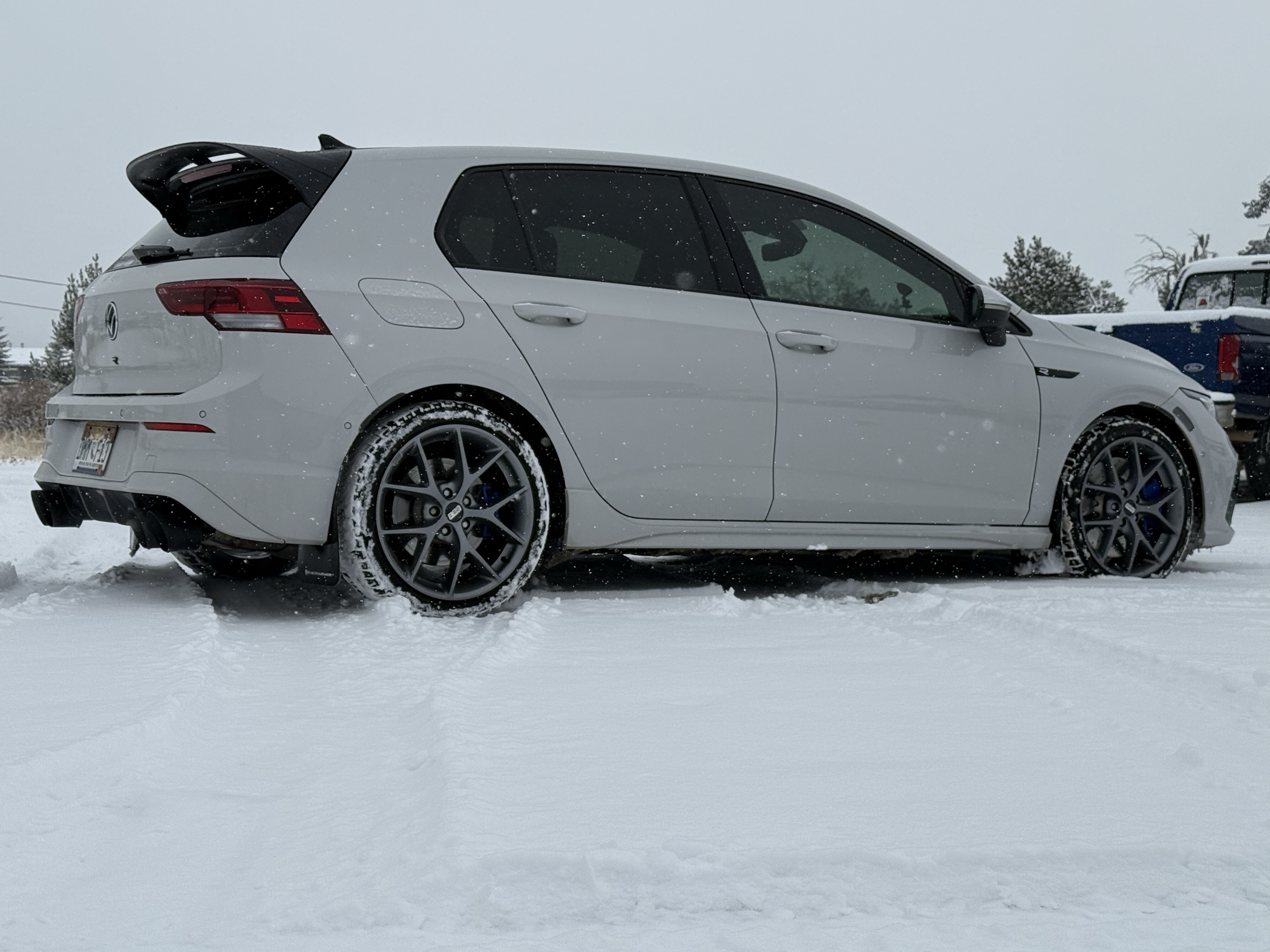 White hatchback car parked in snowy landscape with snow-covered ground and overcast sky.