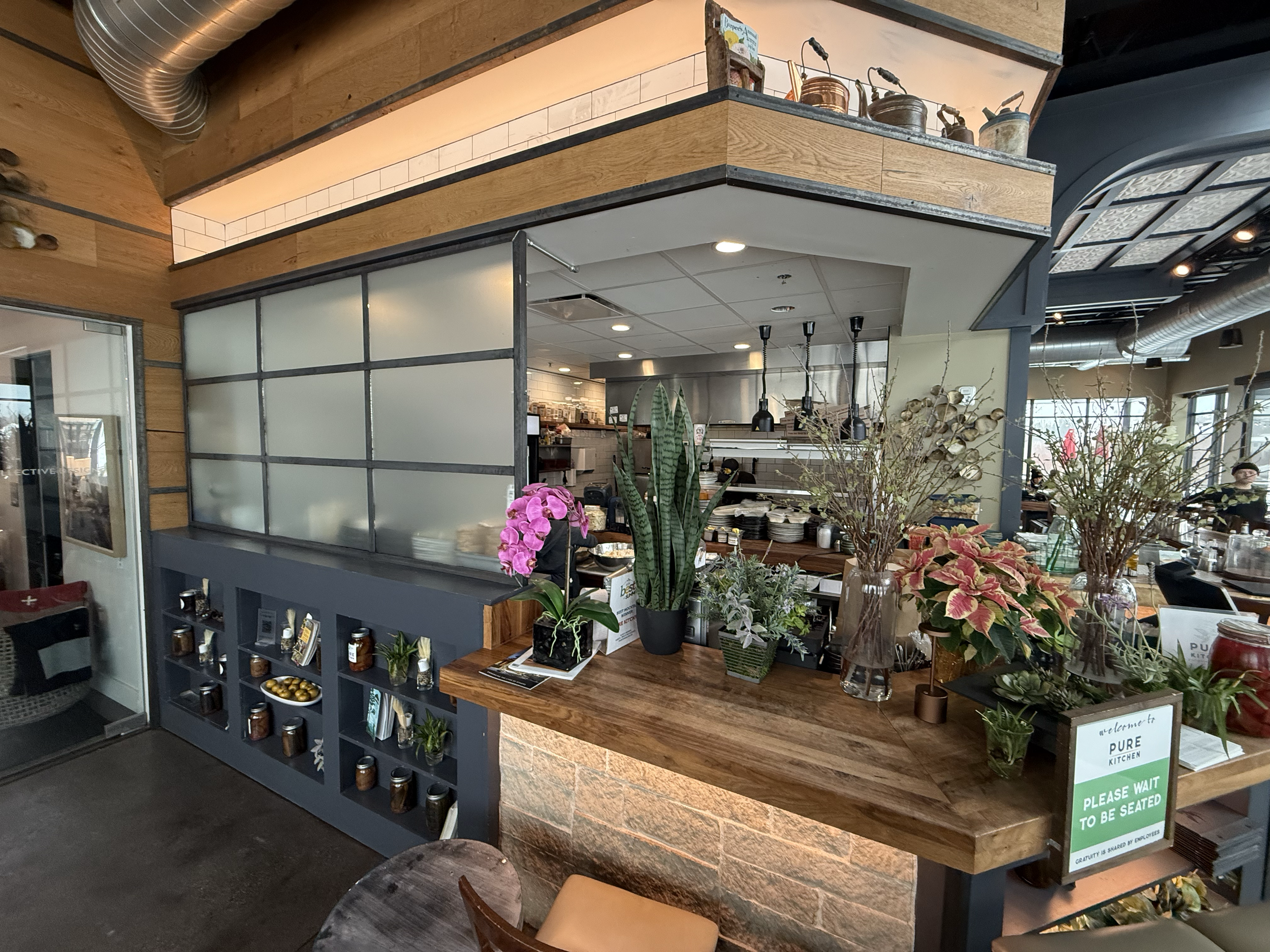 Indoor view of a restaurant's waiting area and kitchen with plants, flowers, and a sign that says 'Please wait to be seated'