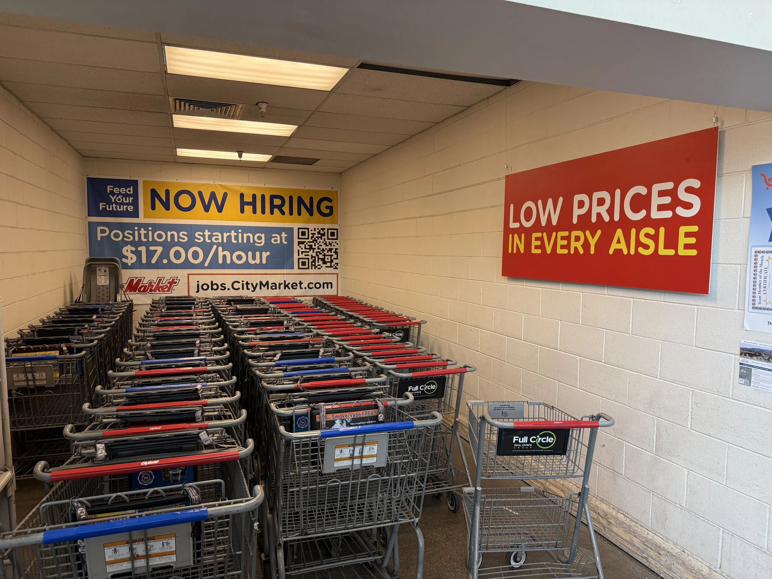 Empty shopping carts in a store aisle with signs advertising now hiring positions starting at $17 per hour and low prices in every aisle.