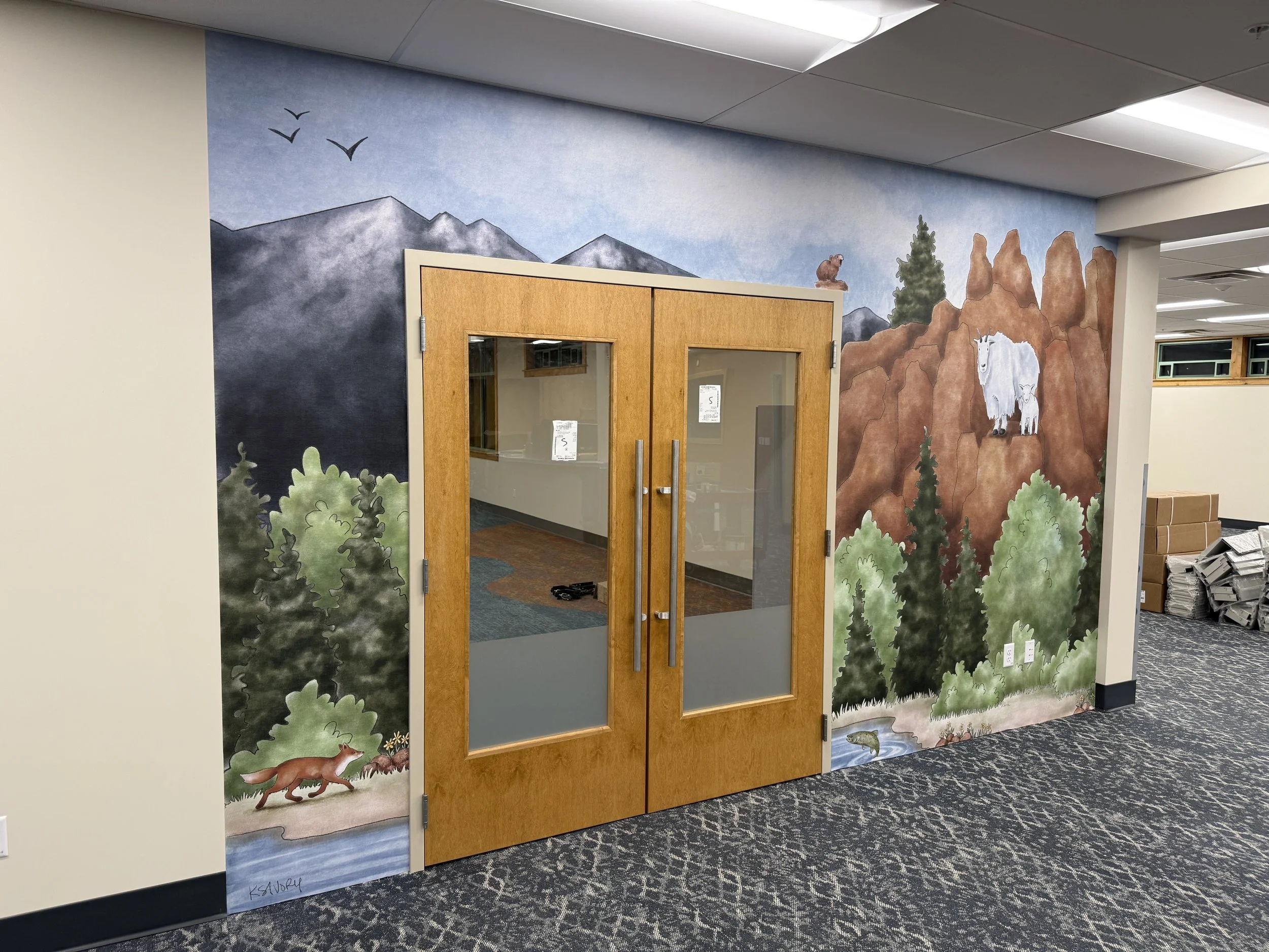 Colorful mural of mountains with birds, a bear, trees, a river, and two mountain goats on a wall behind wooden double doors in an indoor setting.