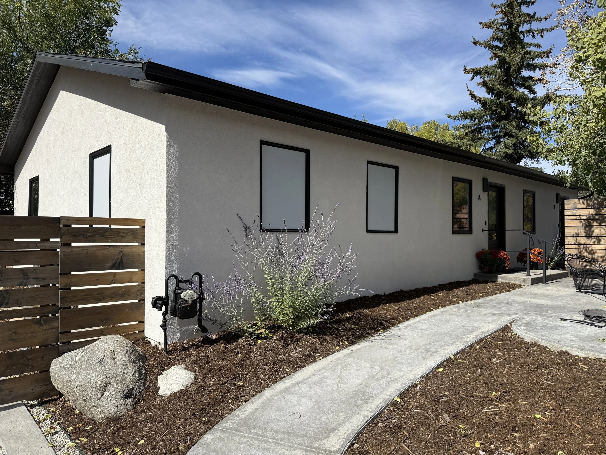 A modern white house with black-framed windows on a sunny day. The front yard has a curved concrete pathway, a small garden bed with mulch, flowering plants, and potted orange flowers. There is a wooden privacy fence on one side and a patio area with