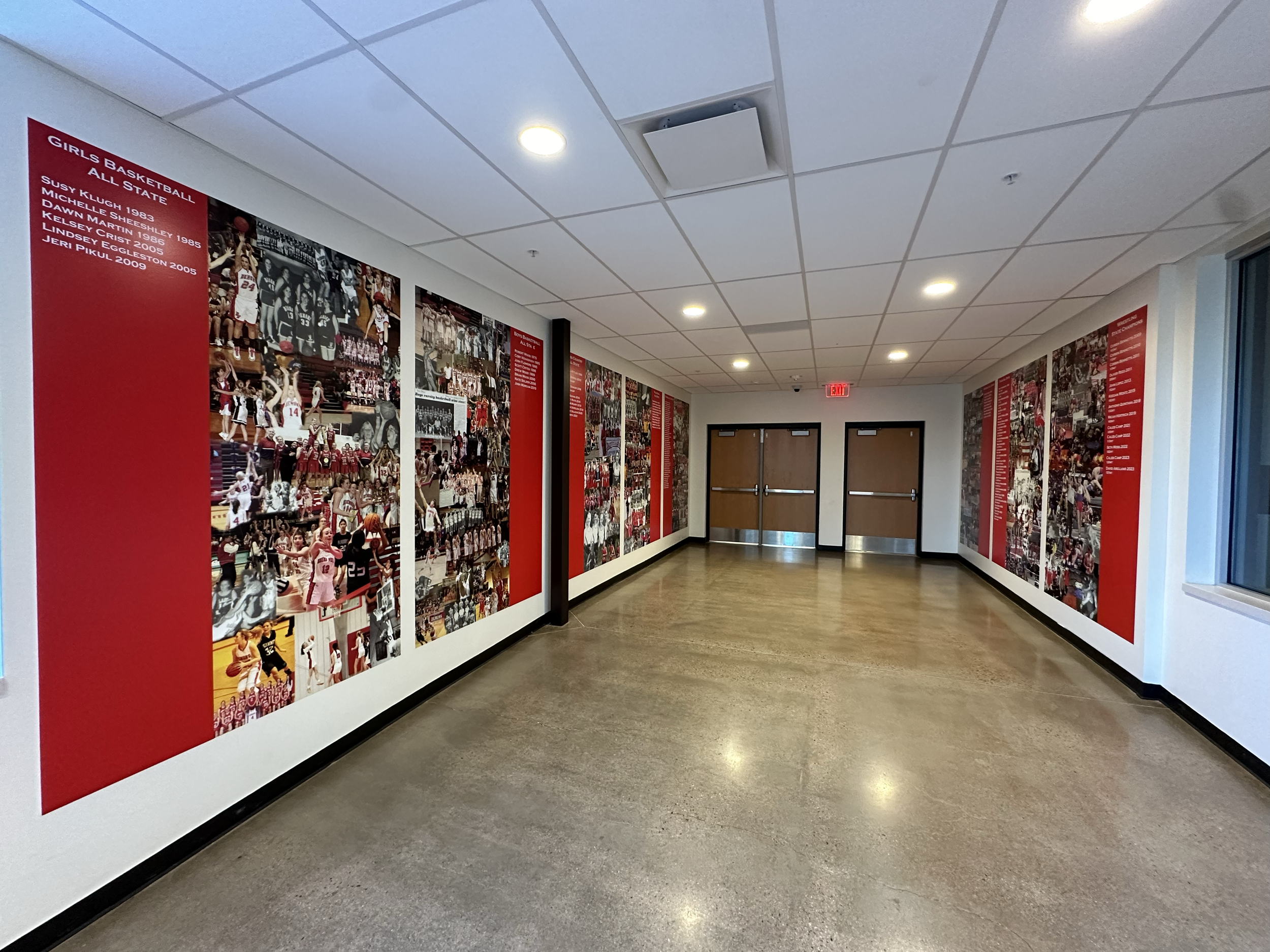 Hallway with basketball team photos and history displayed on red panels along the wall, two closed wooden double doors at the end, exit sign above, and window on the right.