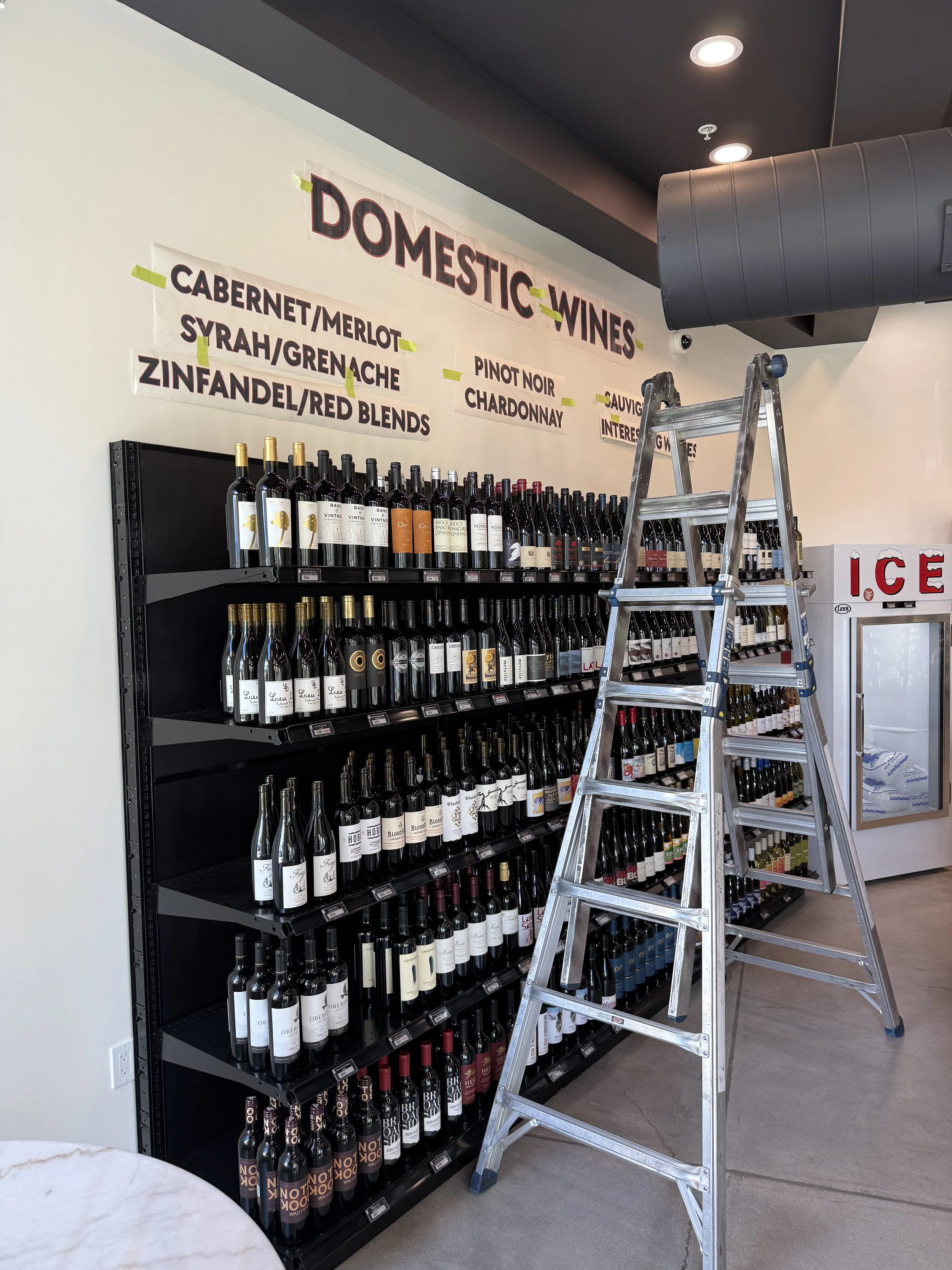 Wall with a sign labeled "Domestic Wines" and variety of wine bottles on shelves, with a ladder in front and a refrigerator labeled "Ice" nearby.