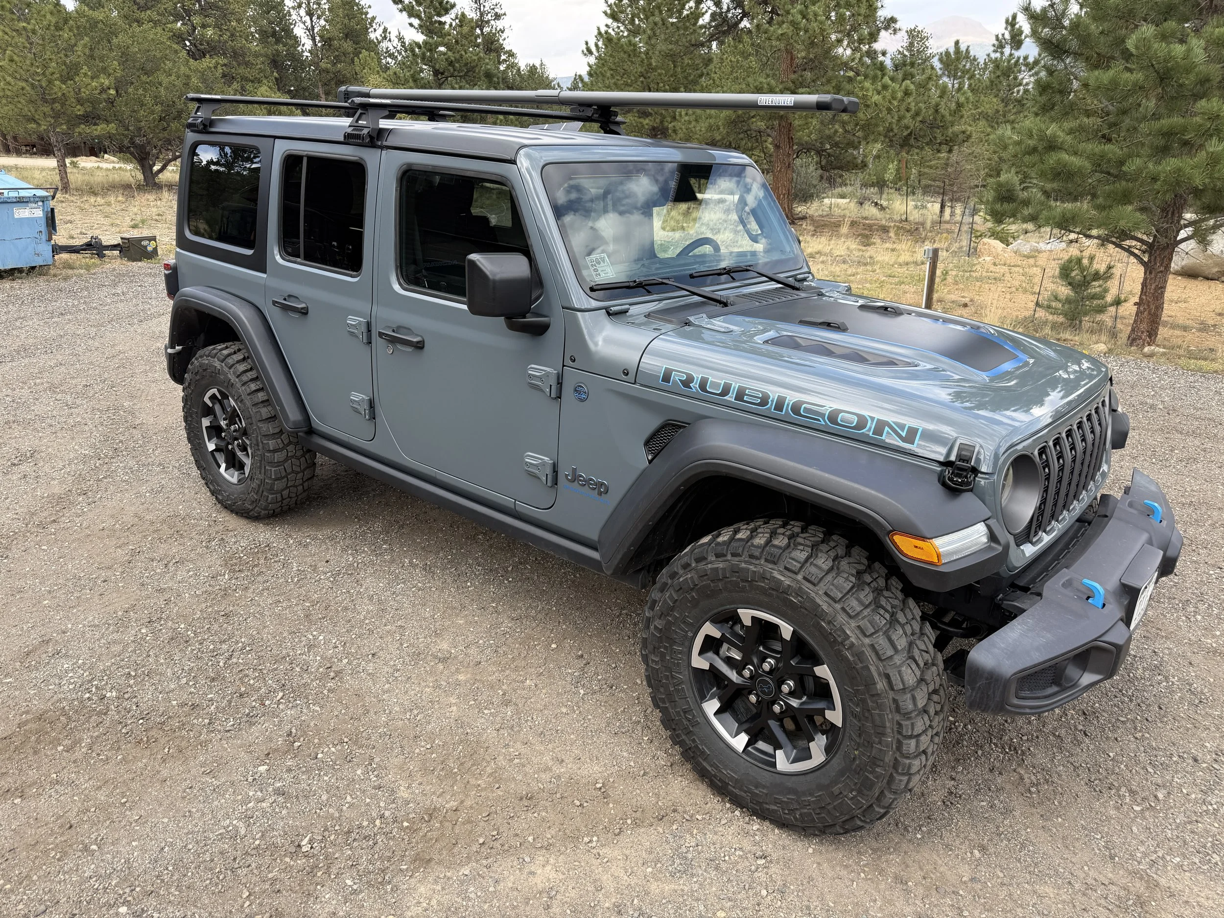 Gray Jeep Rubicon SUV with off-road tires and a roof rack, parked on a gravel surface with trees in the background.