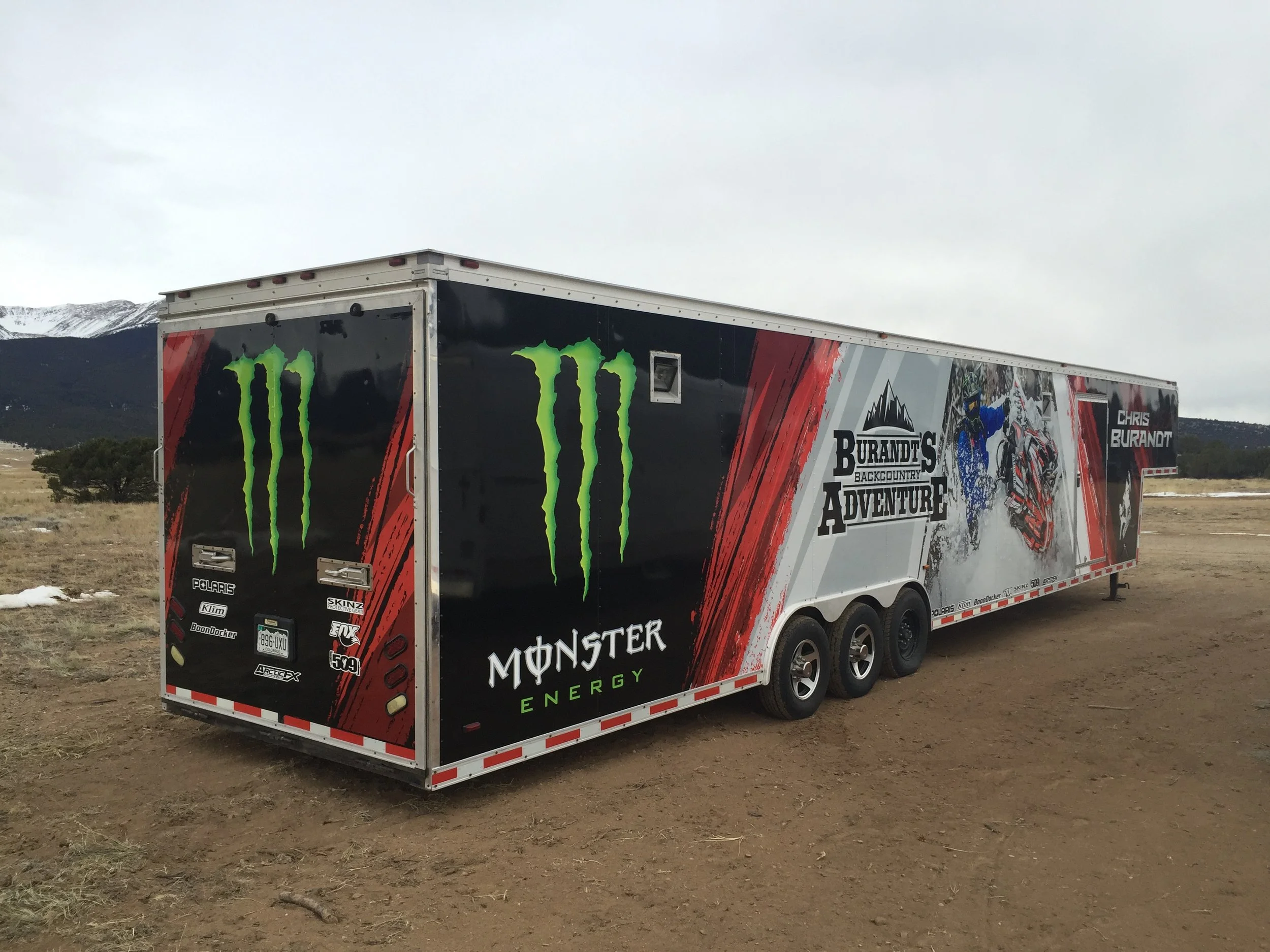 Motocross event trailer with Monster Energy and Chris Burandt branding, featuring a mountain and motocross graphic design, parked on dirt terrain with mountains in the background.