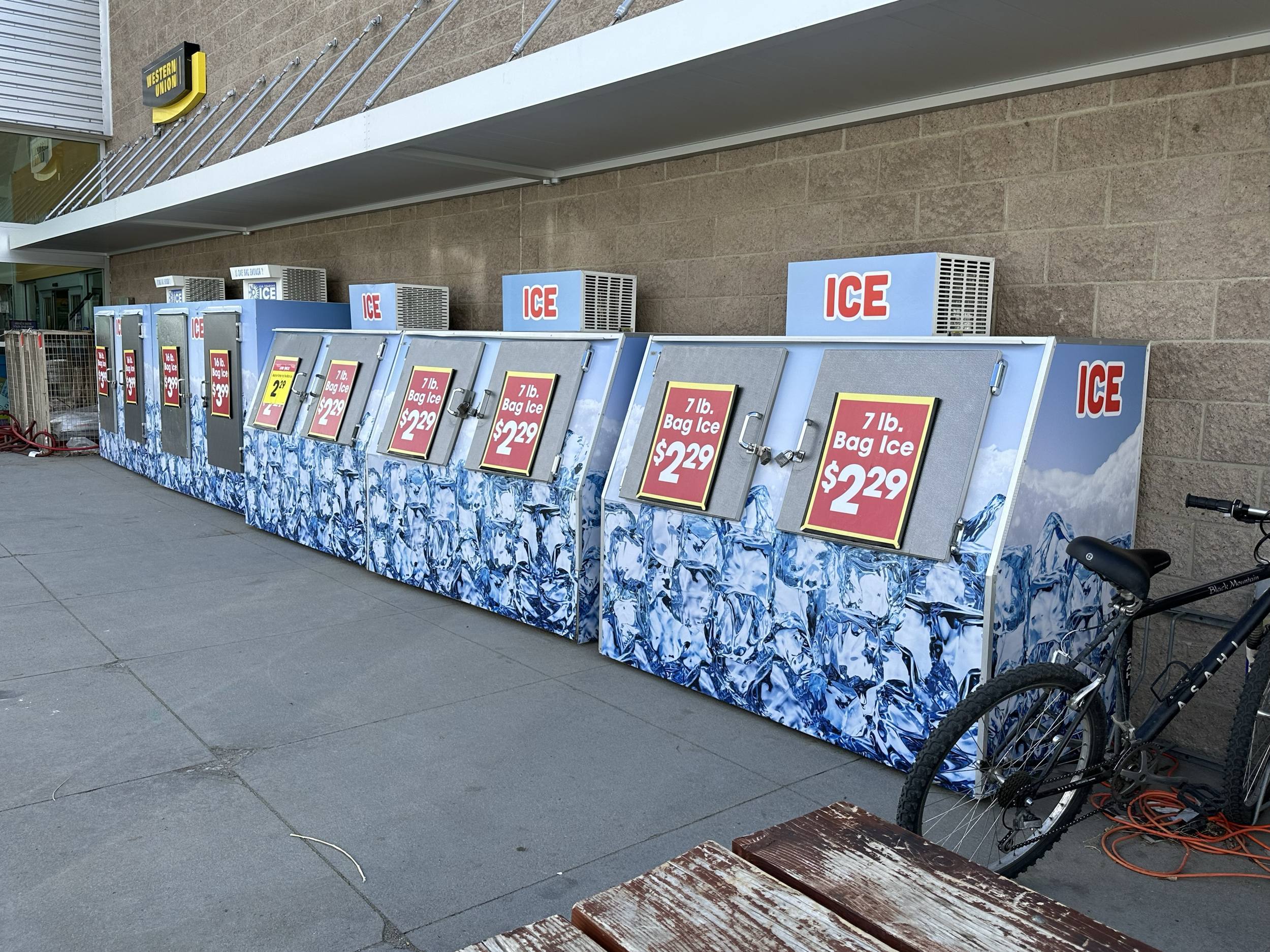 A row of ice vending machines outside a store, each labeled 'ICE,' selling 7 lb. bags of ice for $2.29. A bicycle is parked next to the machines, and the sidewalk is in the foreground.