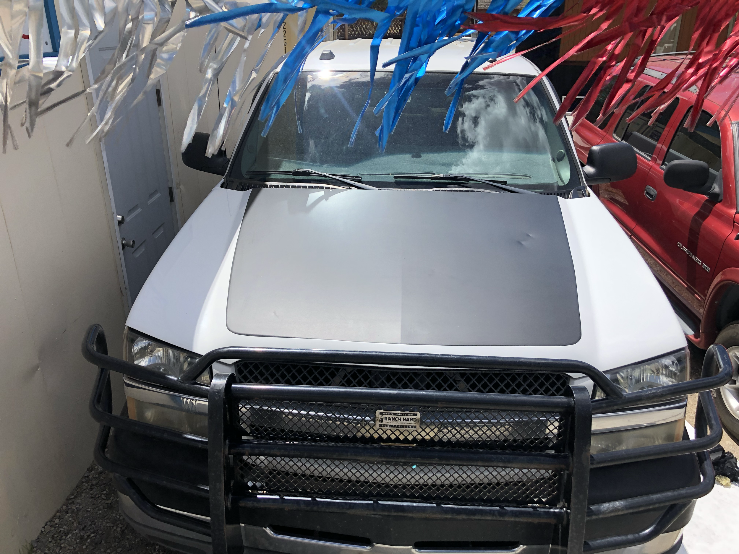 A white pickup truck with a black grill guard parked between a beige building and a red vehicle. The truck has a two-tone hood with a painted black stripe.