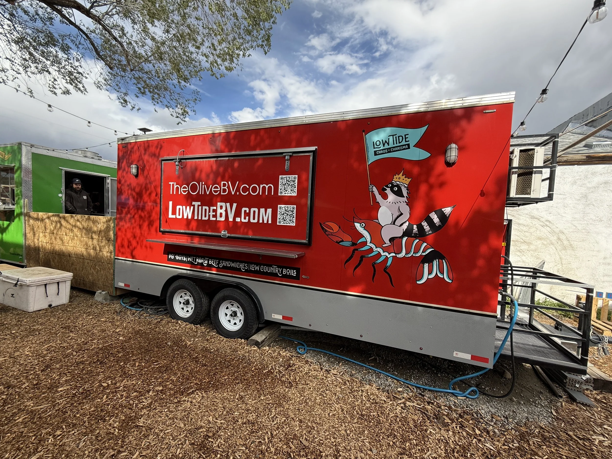 A red food truck with a cartoon raccoon dressed as a king riding a lobster, holding a blue flag that says "Low Tide Chaos & Charisma." The truck has the website "TheOliveBV.com" and "LowTideBV.com" displayed on it, and is parked on a gravel surface u