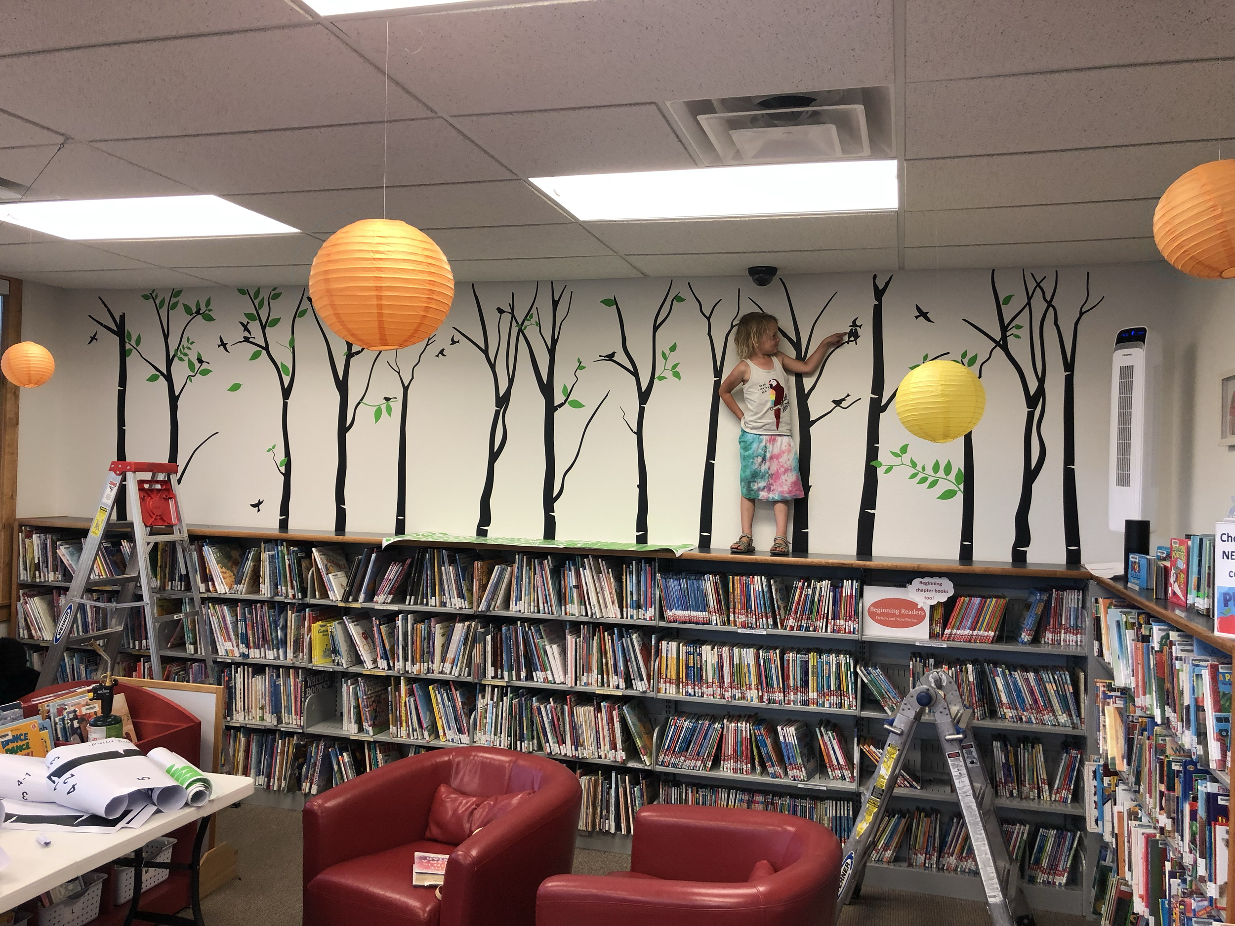 A young girl standing on a ledge inside a library, painting a tree mural on the white wall with black branches and green leaves. There are orange and yellow paper lanterns hanging from the ceiling, and bookshelves filled with books below the mural. T