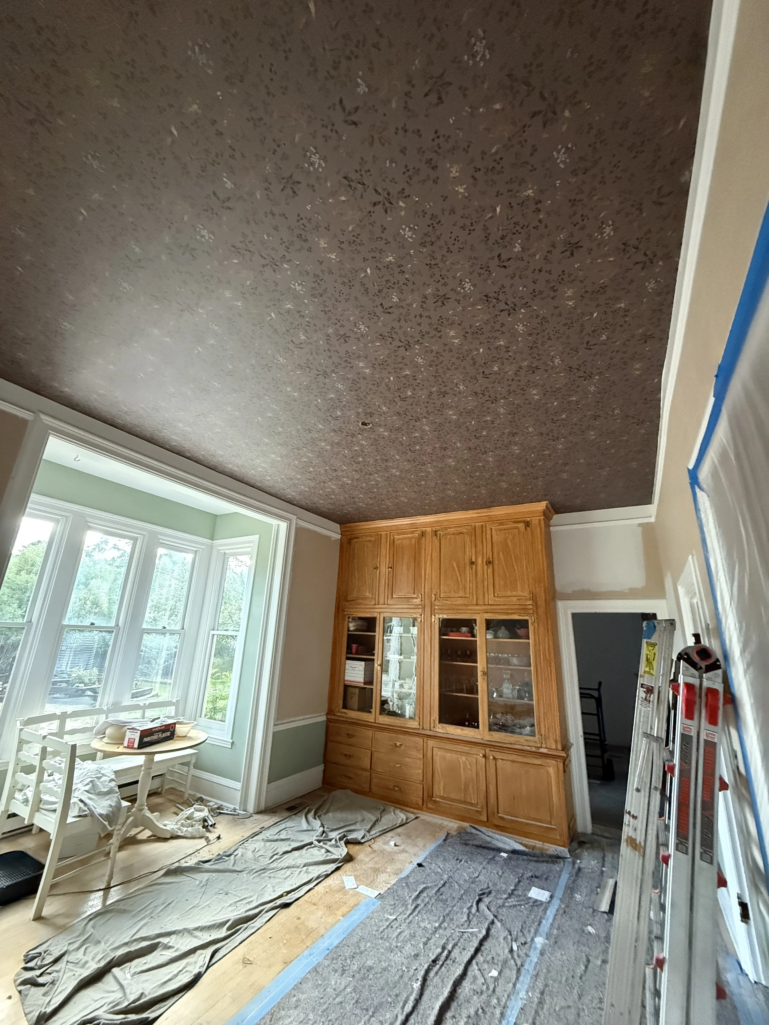 Room with a large window, a wooden built-in cabinet, a ladder, and painting supplies on the floor during renovation.
