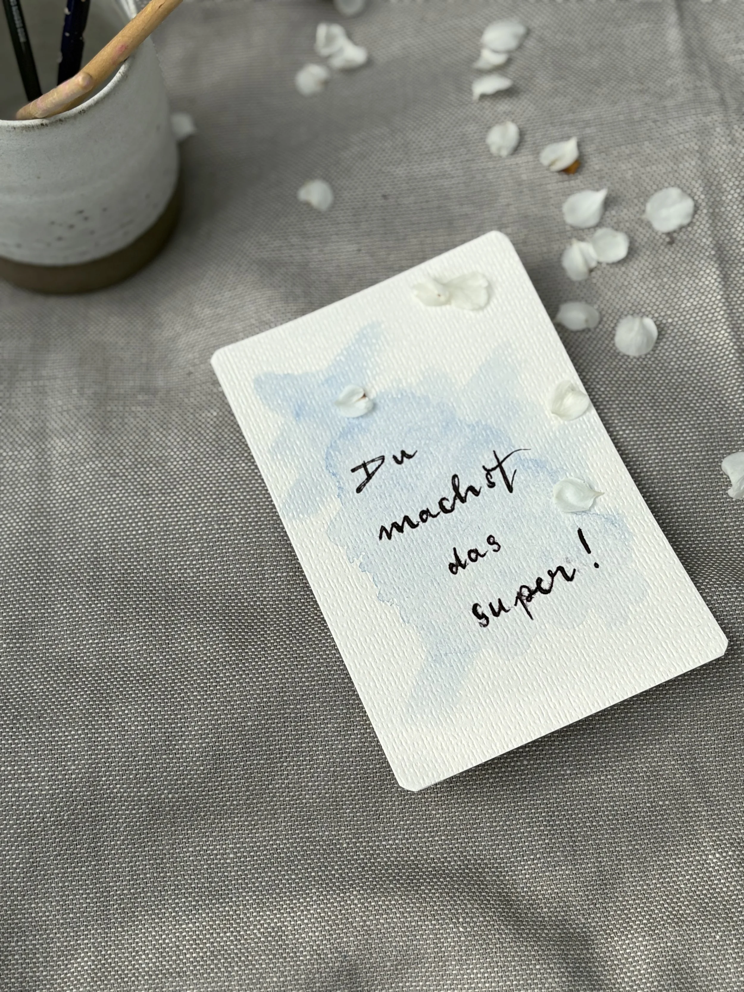 A white card with handwritten text in black ink that says 'Do something today!'. The card is on a gray textured surface. There are scattered white small objects, possibly flower petals or cotton balls, near the card. In the top left corner, there's a gray ceramic container with a wooden stick or brush partially inside.