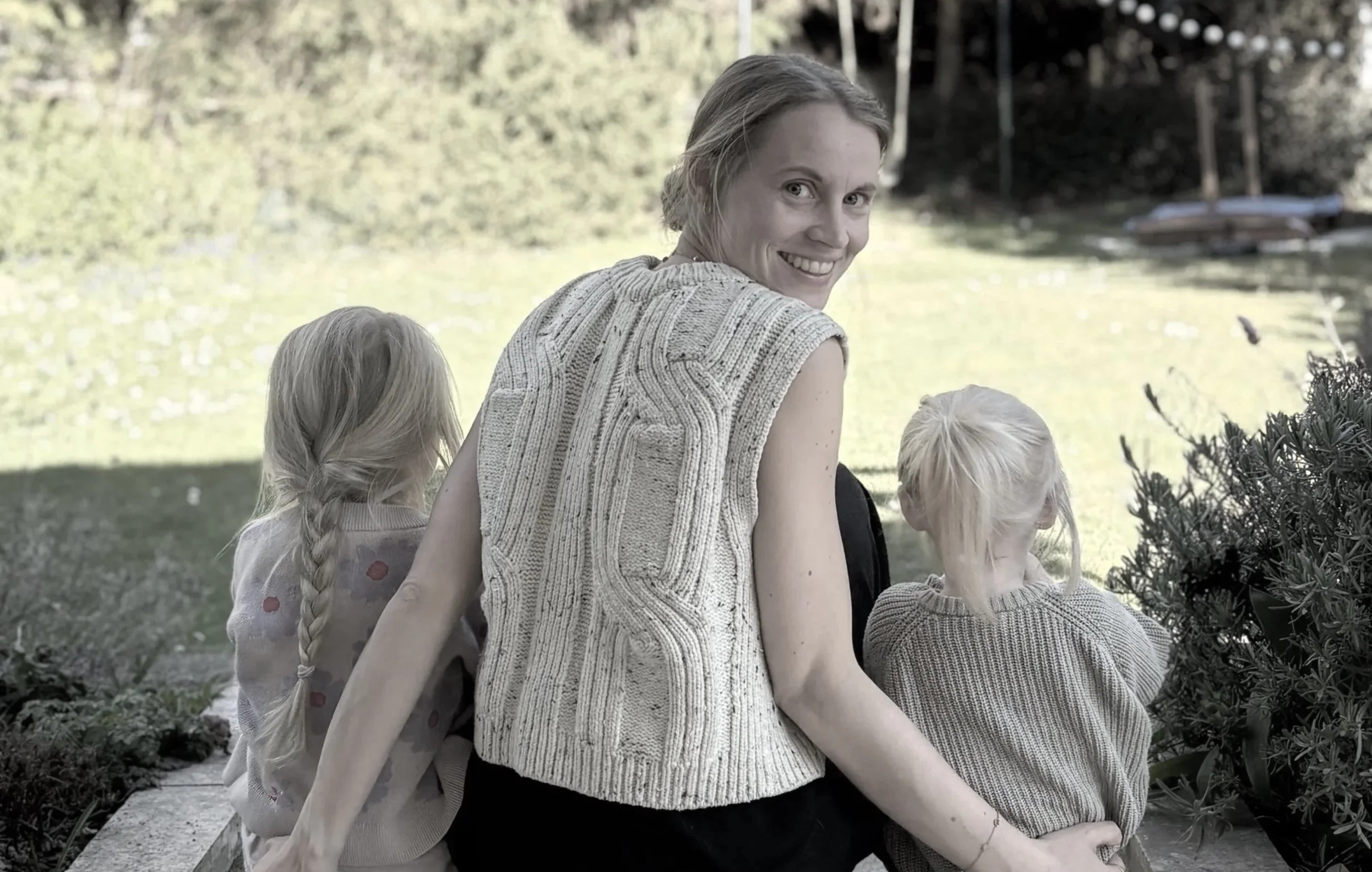 A woman with short, light brown hair smiling and sitting outdoors with three children near a body of water and trees in the background.