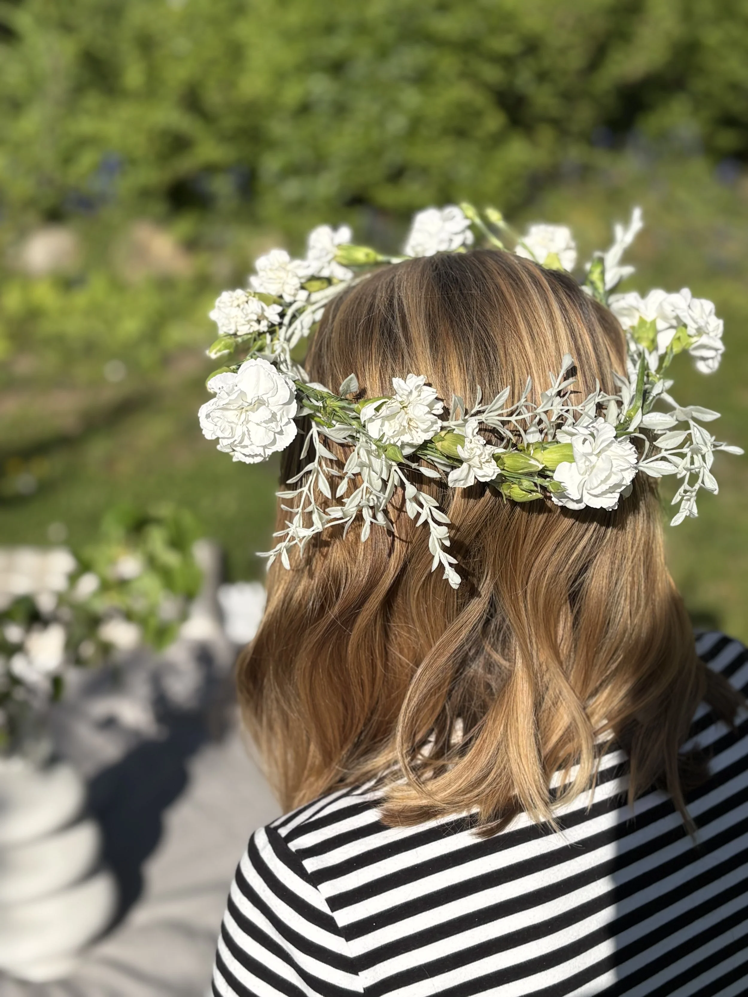 A person with wavy, reddish-brown hair wearing a floral crown made of white flowers, seen from behind. They are outdoors with a blurred green background.