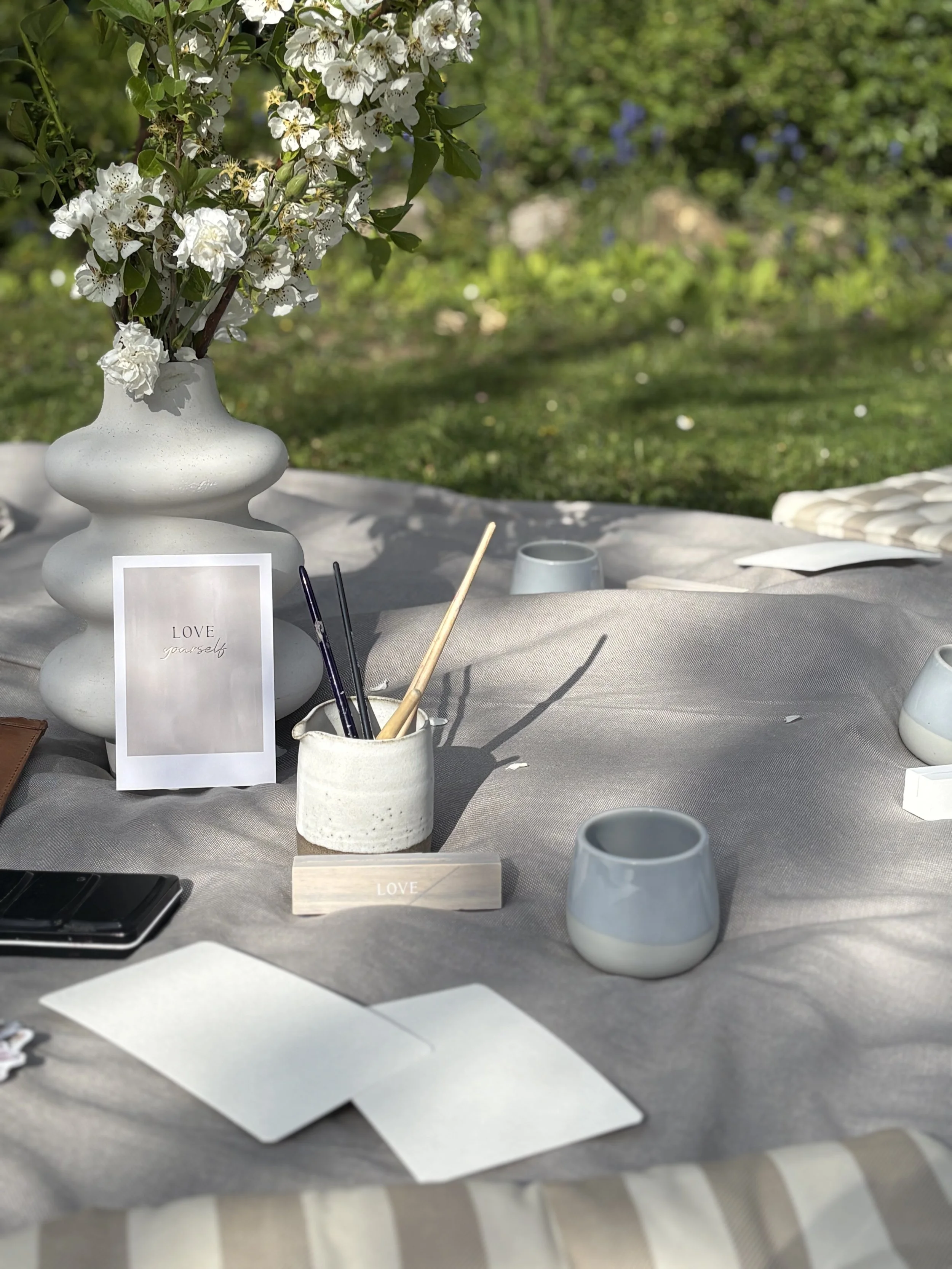A table setup outdoors with a white ceramic vase filled with white flowering branches, a small card with the word 'LOVE' on it, white ceramic cups, and various stationery items on a grey tablecloth.
