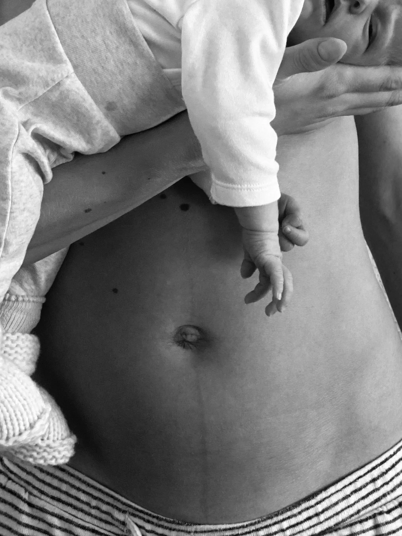 A baby lying on an adult's bare stomach, holding the adult's finger, while the adult covers part of their face with their hand. The baby is dressed in a light shirt, and the adult is shirtless, with the baby on the adult's chest.