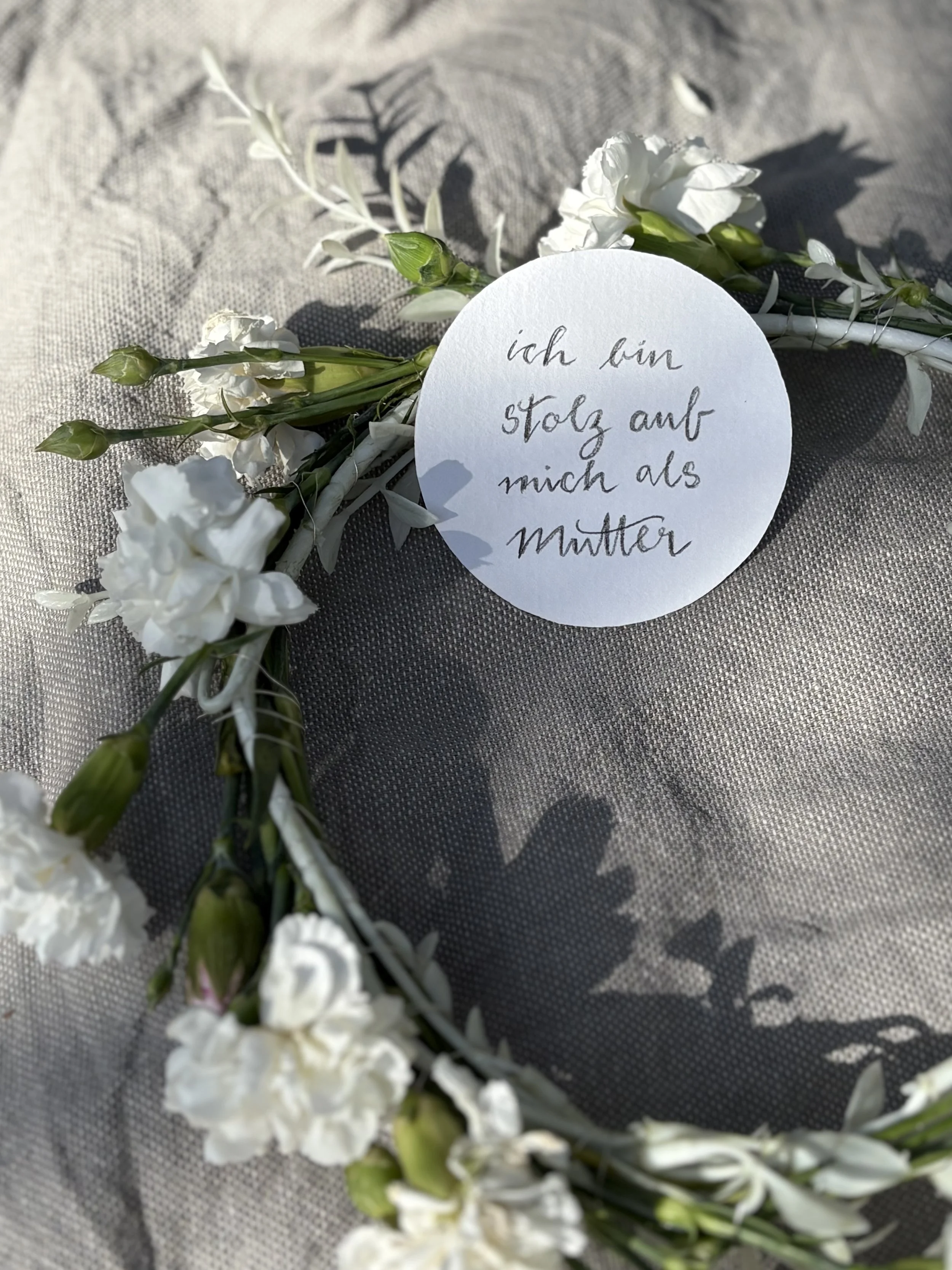 White flower crown with a handwritten note in german on a circular piece of paper, saying 'ich bin stolz auf mich als Mutter'.