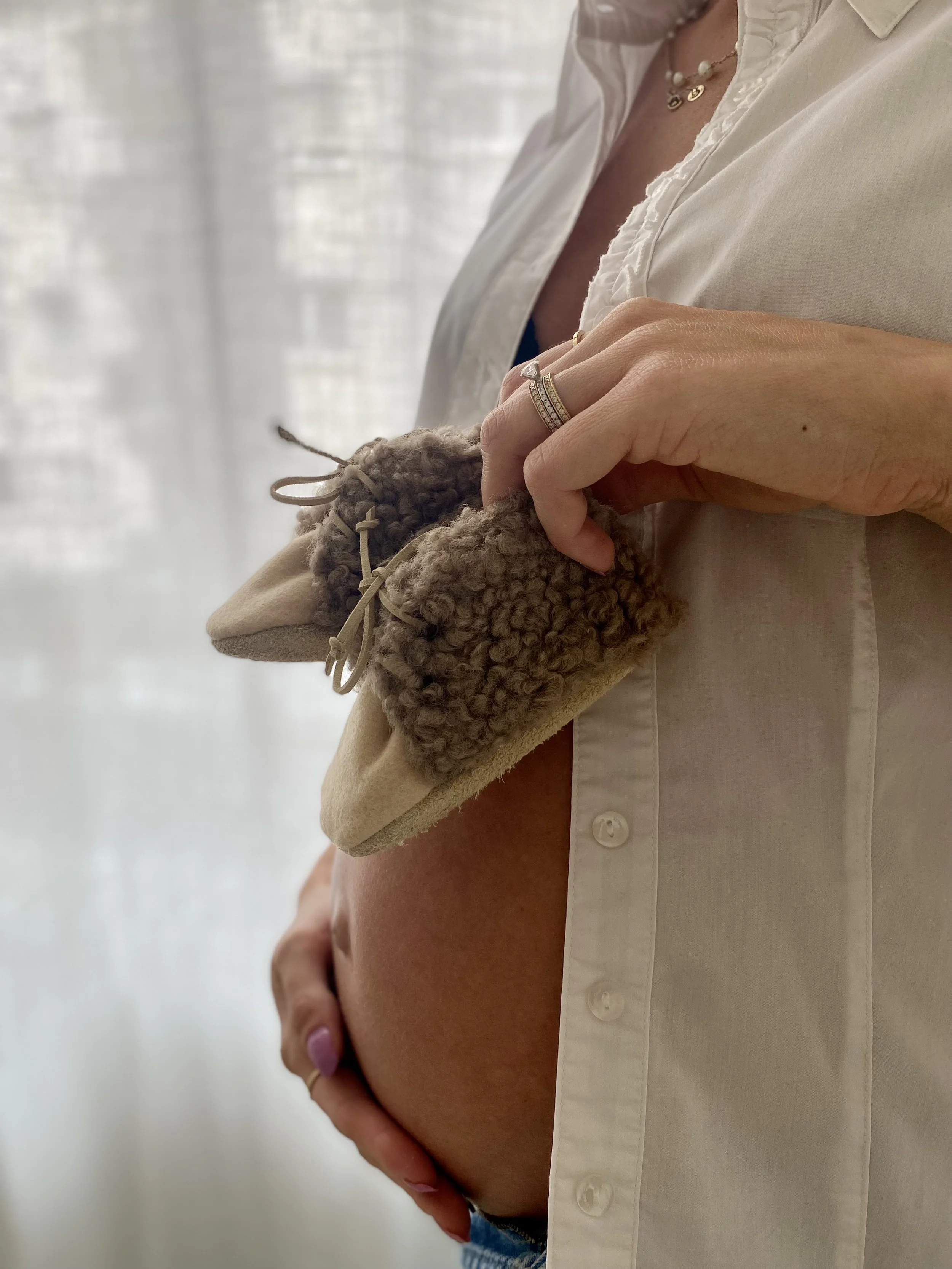 A woman holding her pregnant belly with her right hand, wearing multiple rings and a beige shirt with buttons, standing in front of a sheer curtain.
