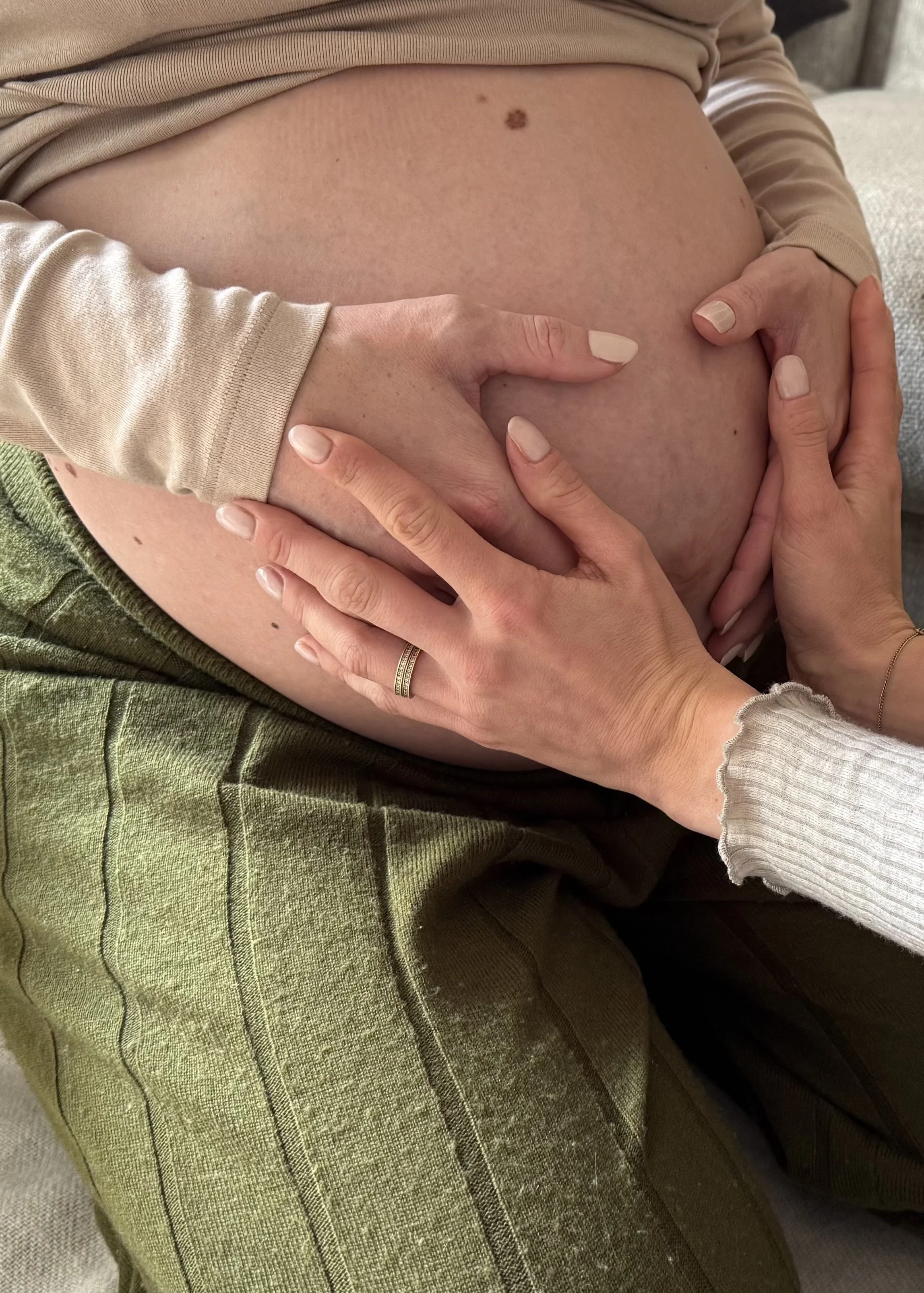 Person's hand gently touching a pregnant belly, wearing a beige long sleeve shirt, specific focus on the belly and hands.