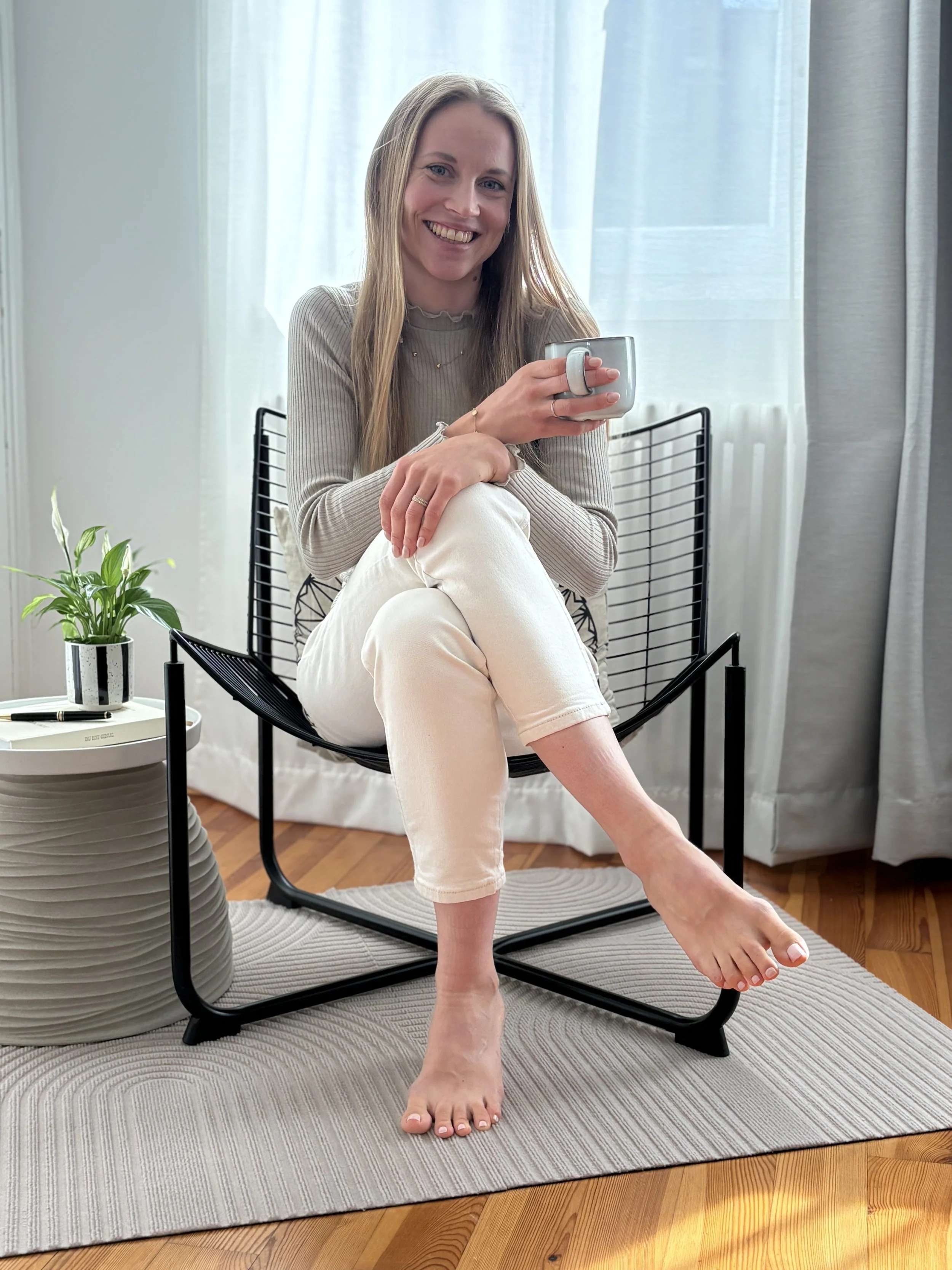 A woman sitting on a black wire chair in a bright room, holding a mug, smiling, with a potted plant nearby, and a white curtain in the background.