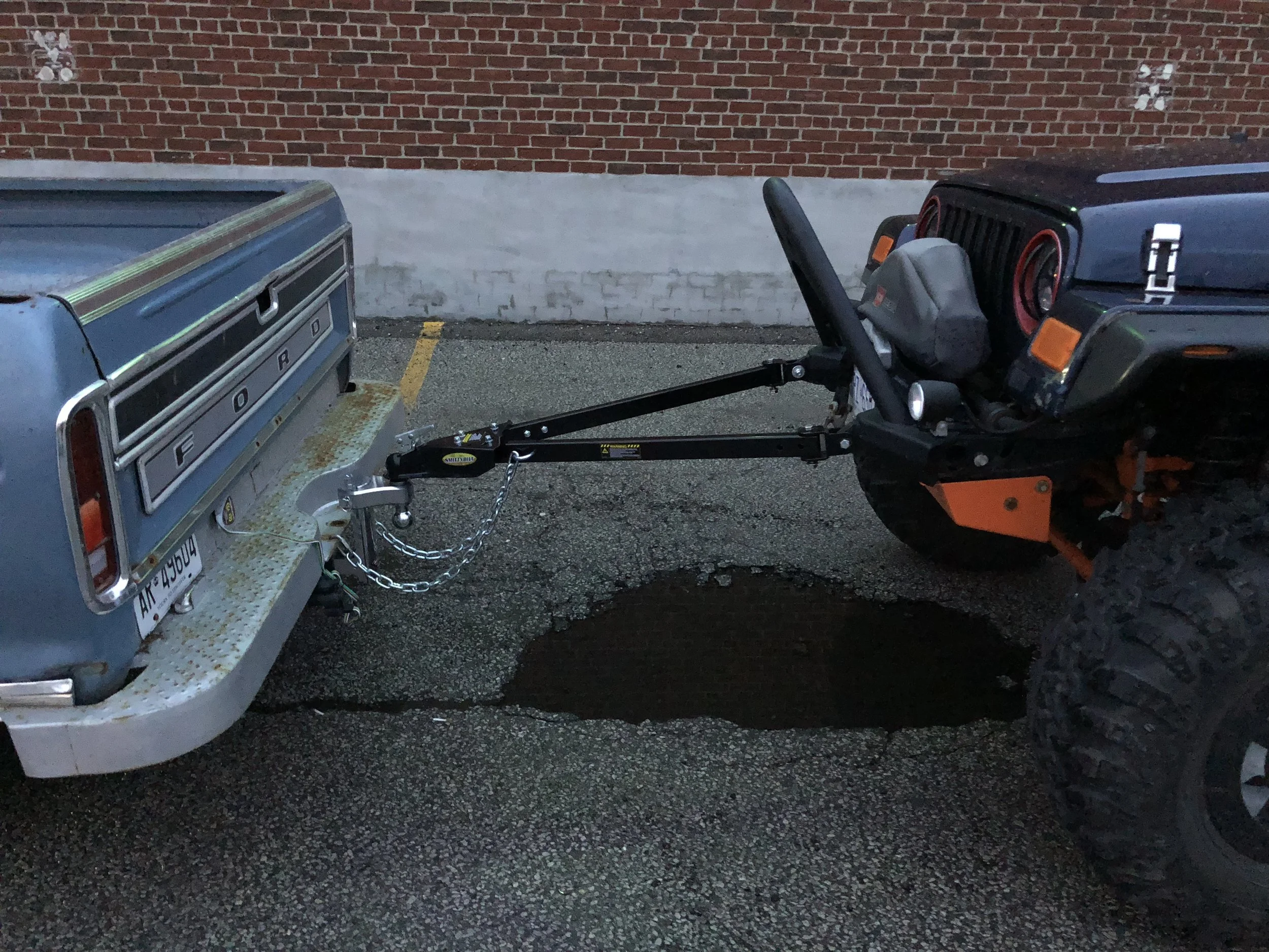 Jeep being towed with a tow bar behind a Ford pickup in a parking lot.