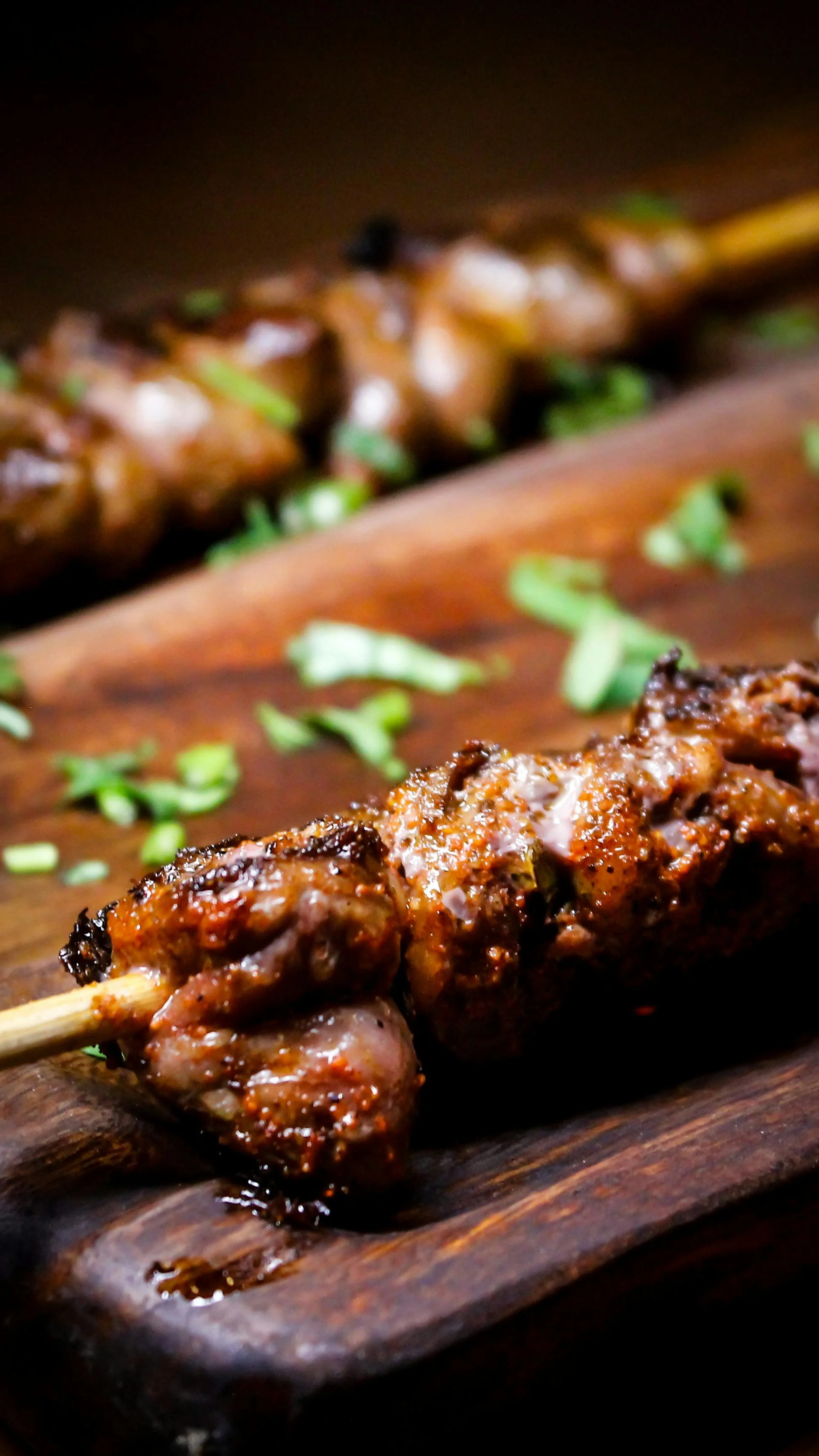 Close-up of grilled meat skewer on a wooden serving board, garnished with chopped green herbs.