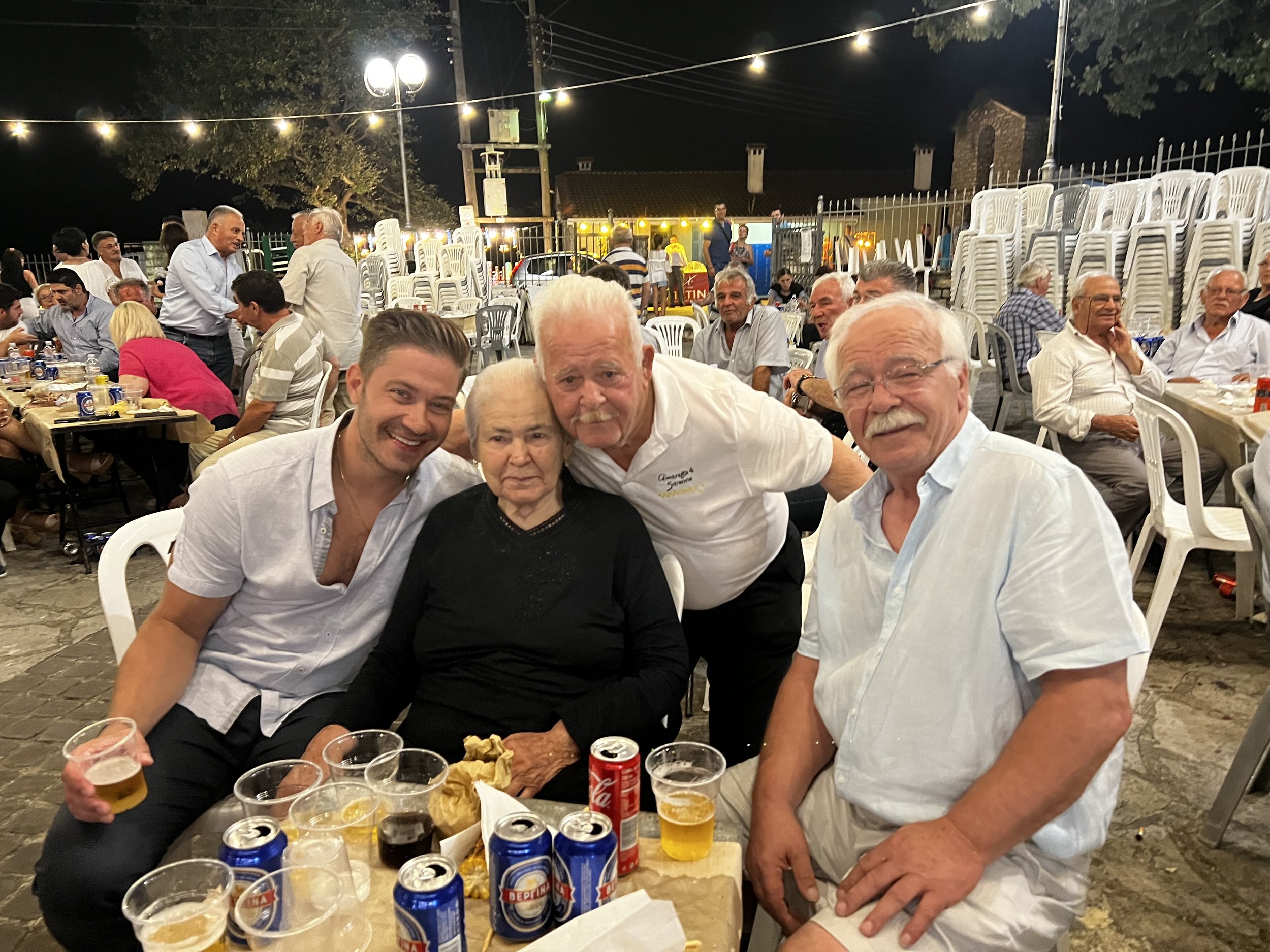 A group of people gathered at a nighttime outdoor event, sitting and standing around tables with drinks and food, under string lights with a fence and trees in the background.